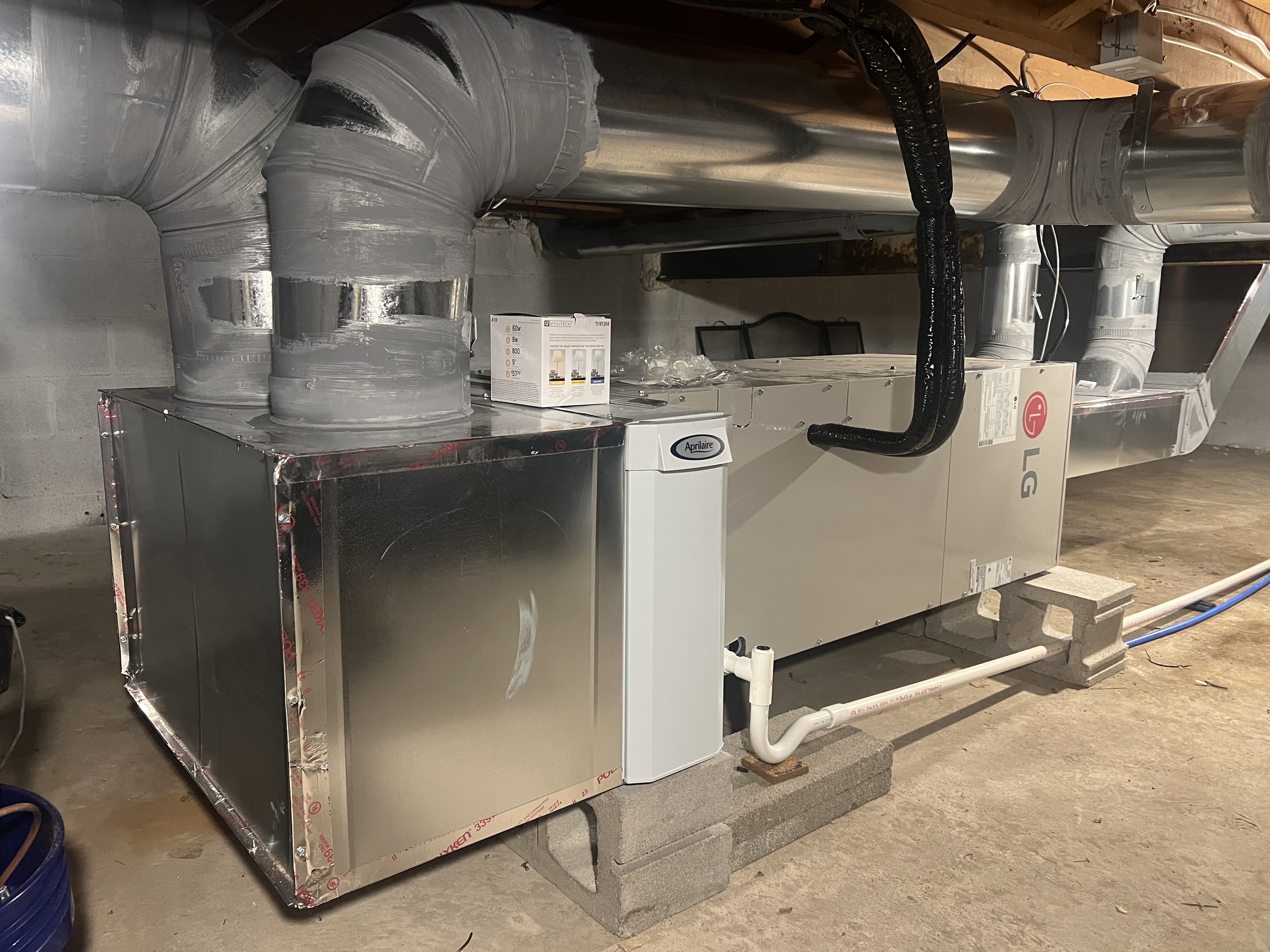 Indoor HVAC unit installed in a basement, featuring metal ductwork and an Aprilaire device. The setup includes pipes, sealing tape, and cement blocks for support.