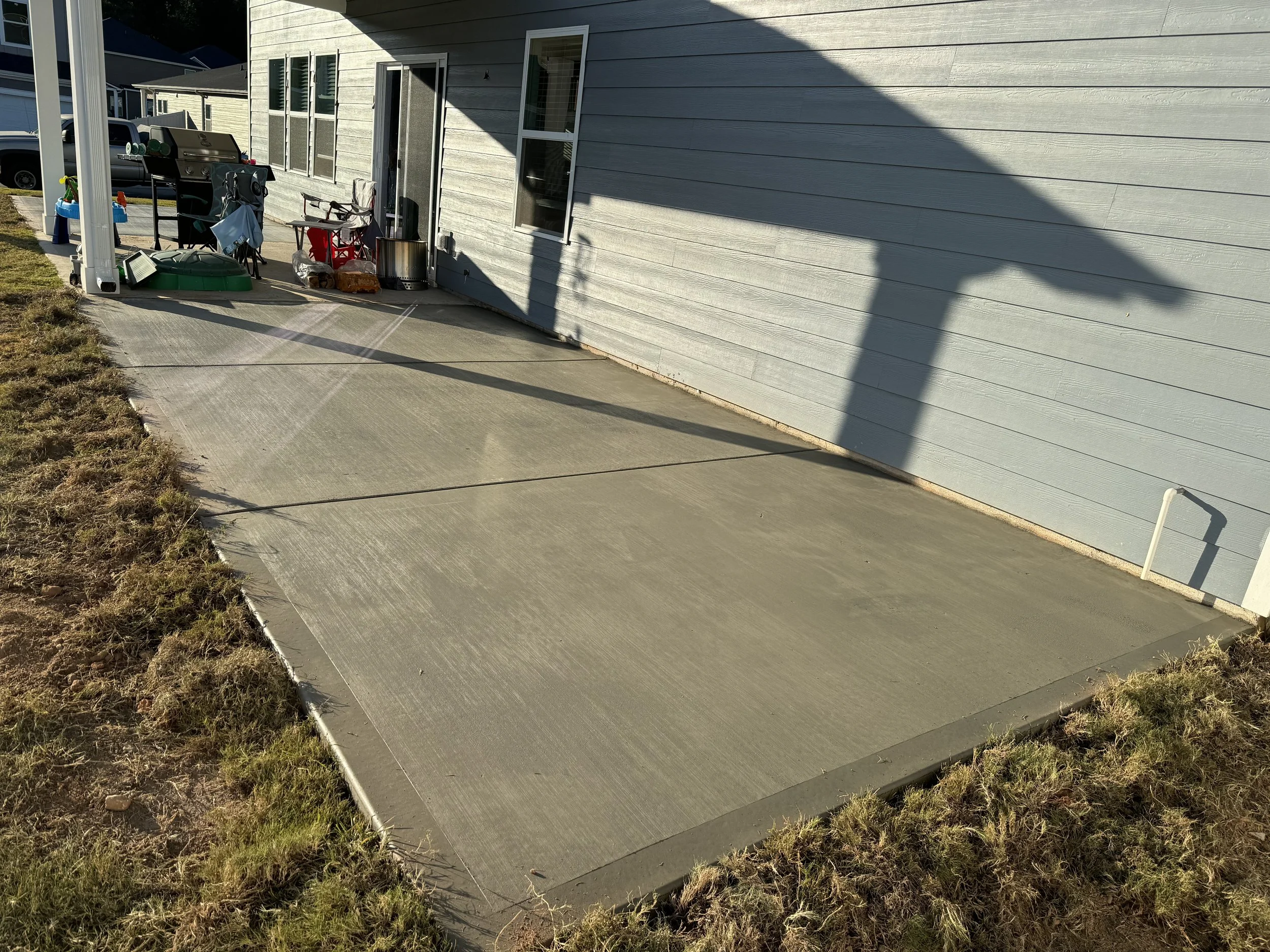 Newly poured concrete patio next to a house with light gray siding, with supplies and tools on the patio and a shadow cast from a covered porch.