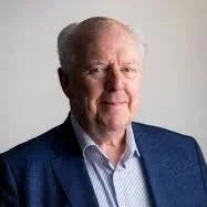 Portrait of an elderly man with white hair, wearing a blue blazer and a light-colored collared shirt, against a plain light background.