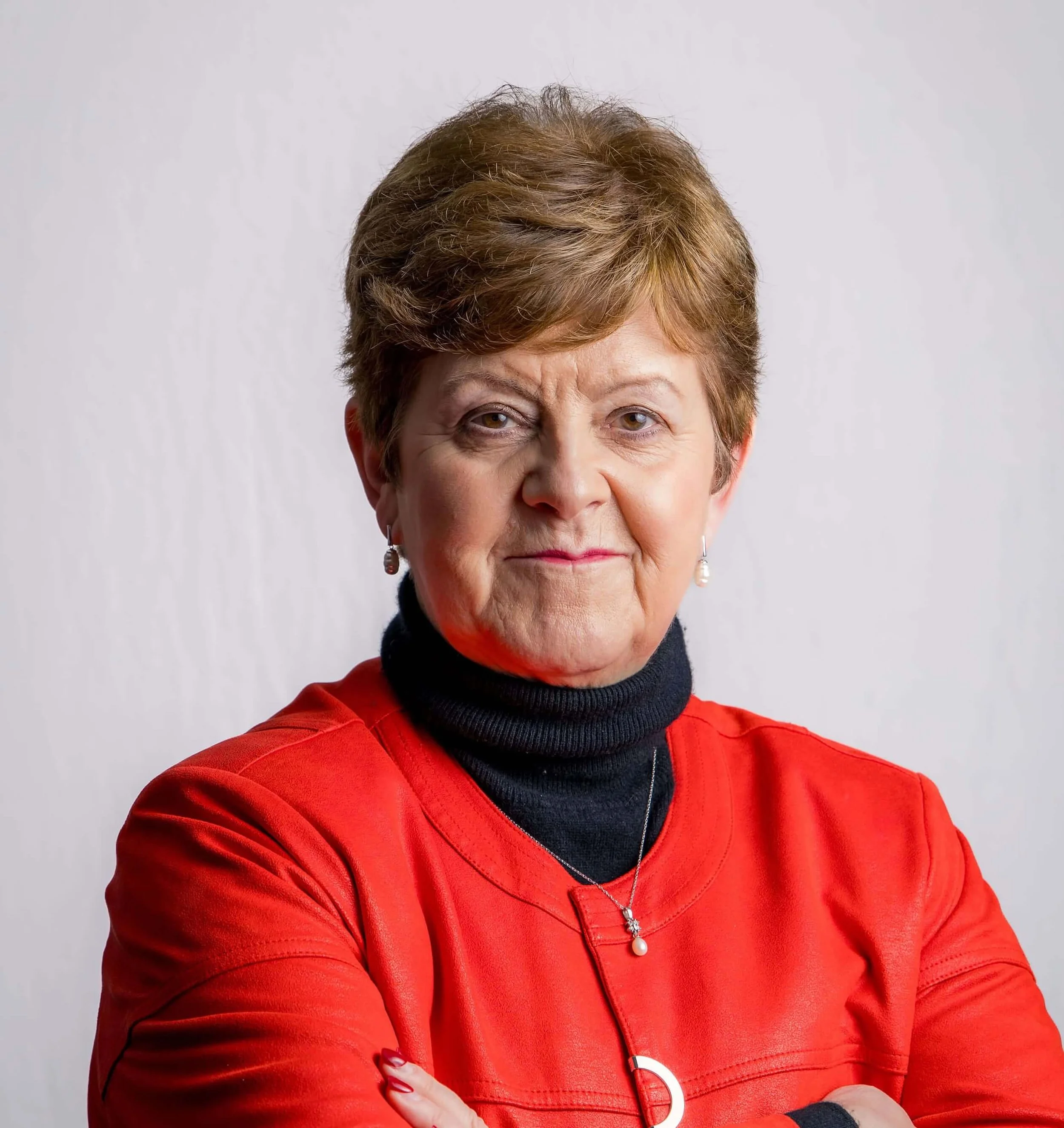 Close-up of a middle-aged woman with short, light brown hair, wearing a red jacket over a black turtleneck, standing against a plain light gray background.