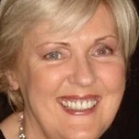Close-up of a smiling middle-aged woman with blonde hair and light-colored eyes, wearing a pearl necklace.