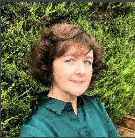 A woman with curly brown hair and light skin, wearing a teal blouse, standing in front of green shrubs.