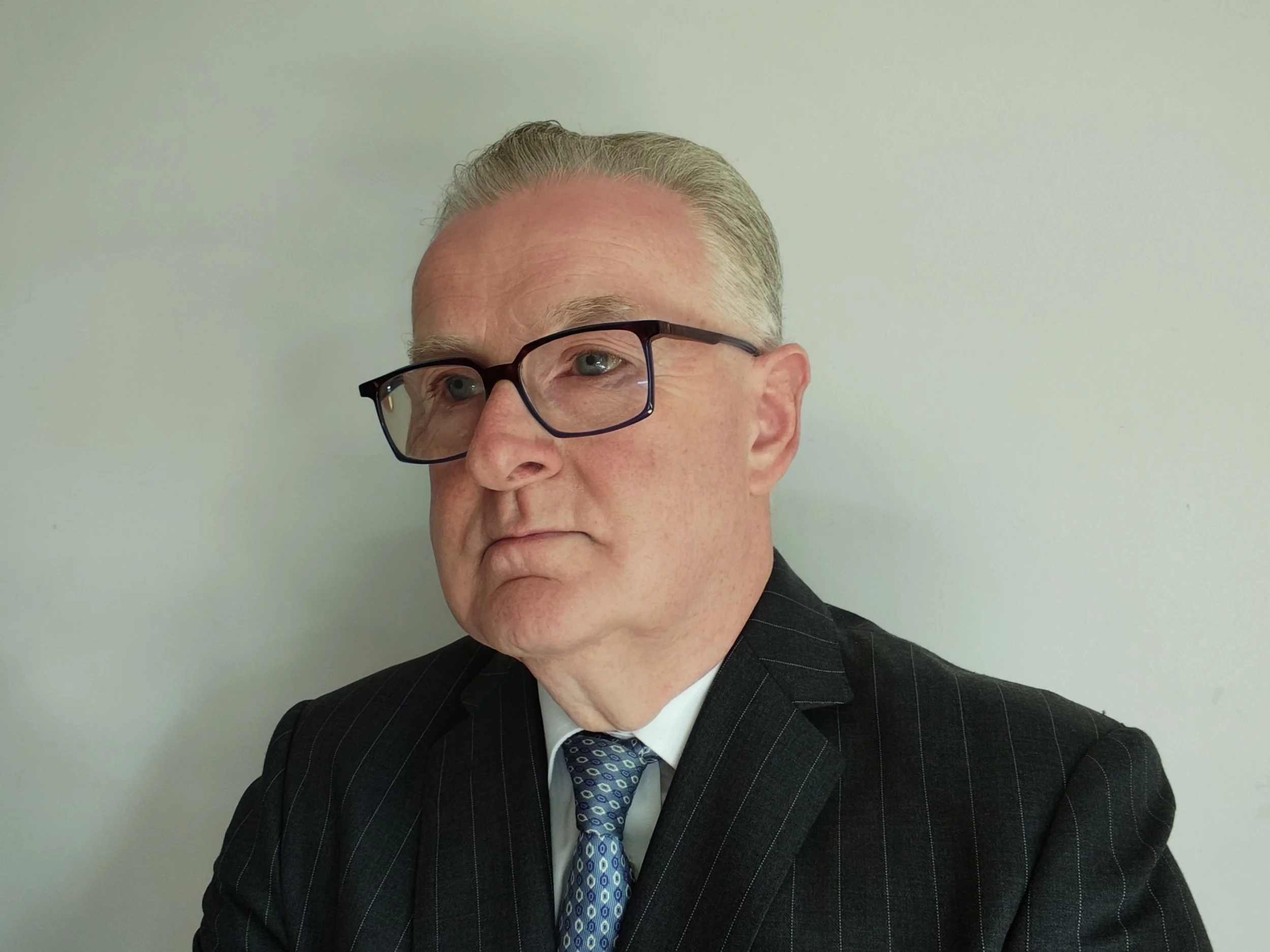 Close-up of a middle-aged man with gray hair, glasses, wearing a dark pinstripe suit, white shirt, and a blue patterned tie, against a plain light-colored wall.
