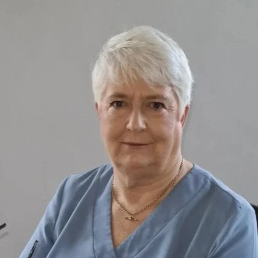 A woman with short white hair wearing light blue scrubs and a gold necklace, sitting against a plain wall.