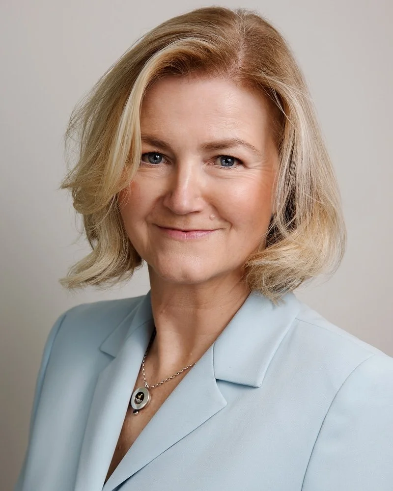 A professional woman with blonde hair, blue eyes, and fair skin smiling, wearing a light blue blazer and a necklace, standing against a neutral background.