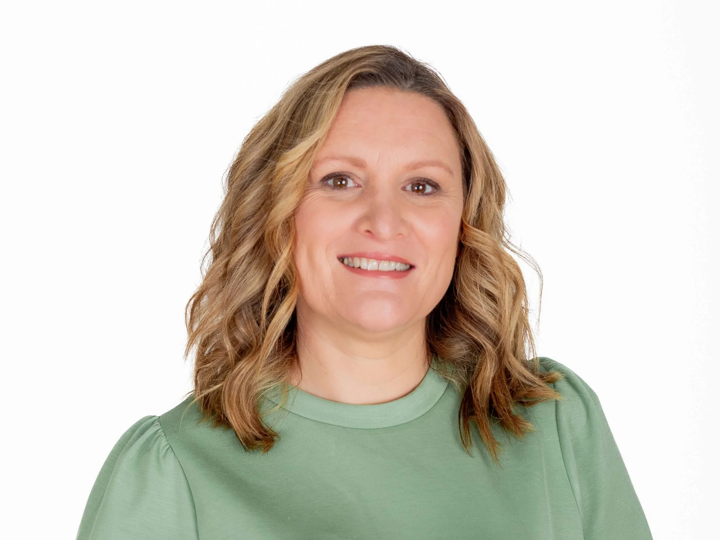 A woman with shoulder-length wavy blonde hair wearing a green dress or top, smiling against a plain white background.