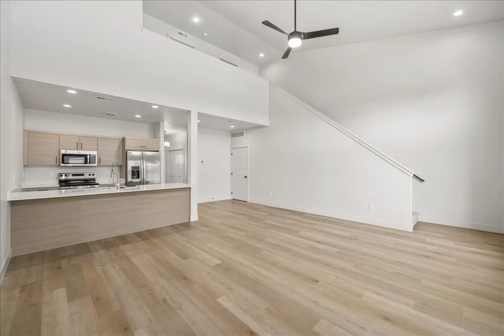 Open-concept living space with kitchen and loft, featuring light wood floors, white walls, ceiling fan, and recessed lighting.