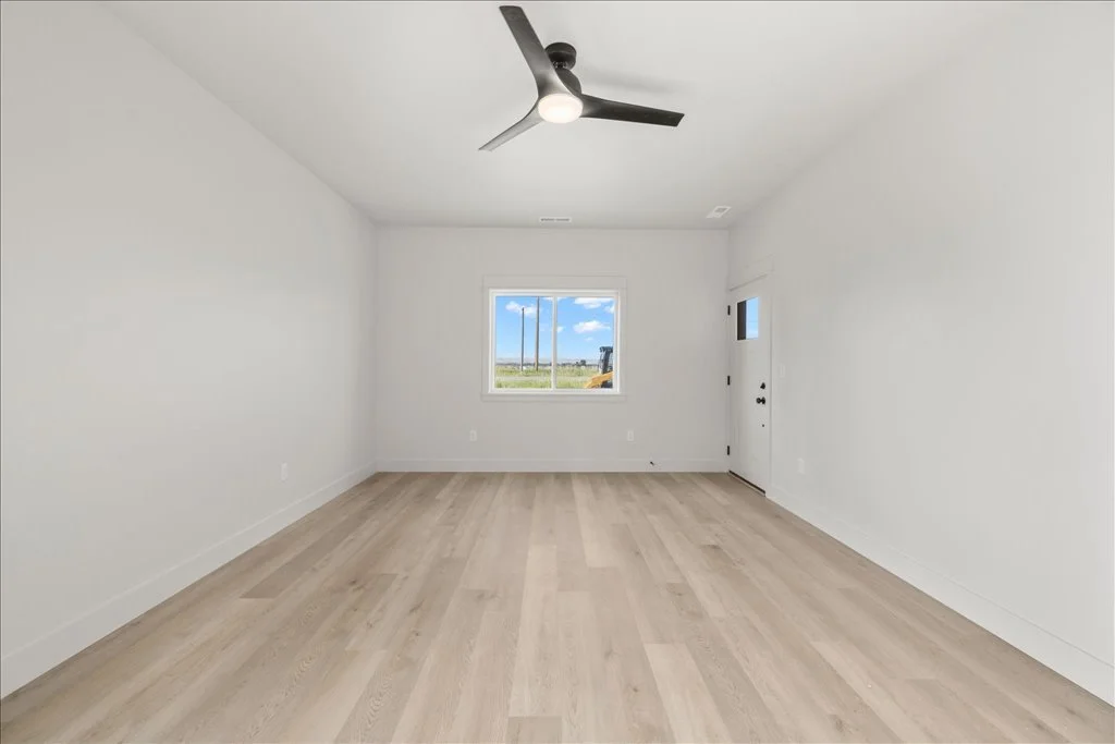 Empty room with white walls, a window showing a blue sky with clouds, a ceiling fan, and light-colored wood flooring.