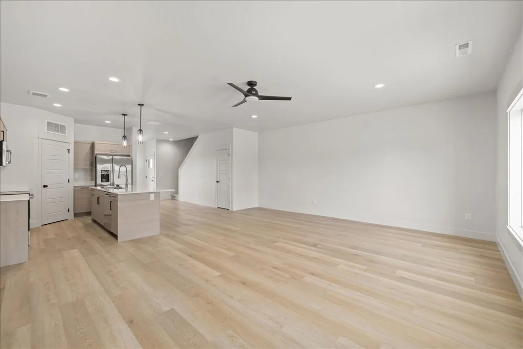 Empty open-concept living room and kitchen with light wood flooring, white walls, and large windows.