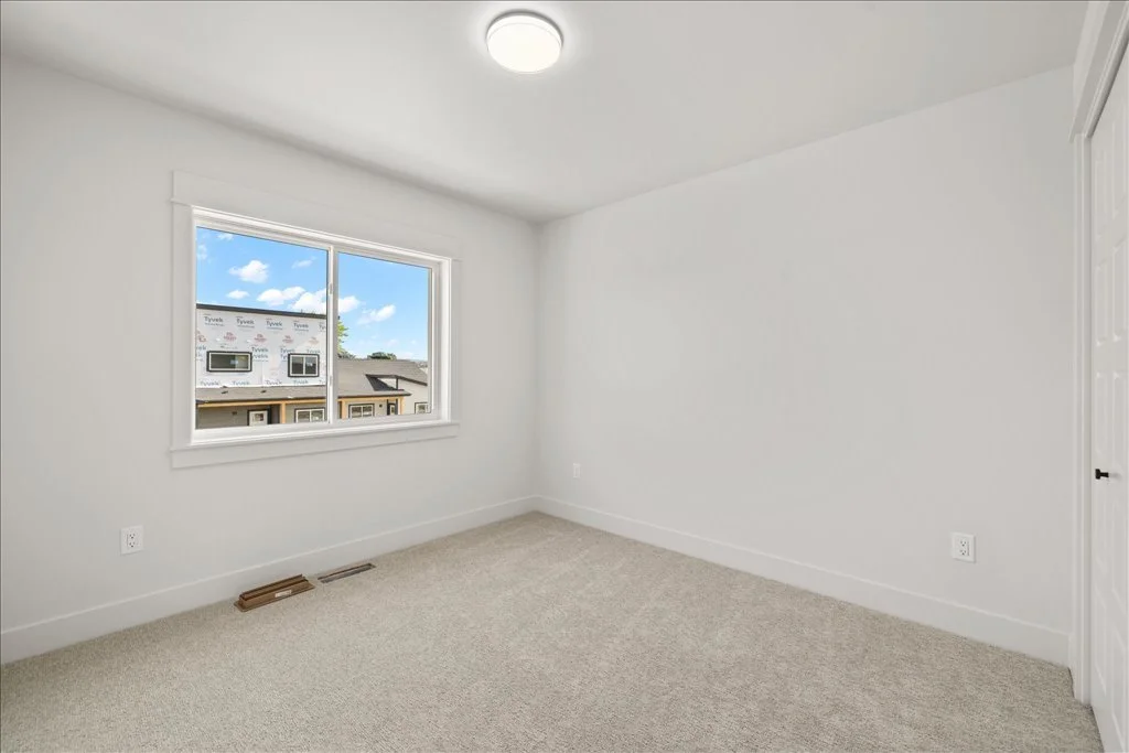 Empty white bedroom with window showing houses and blue sky with clouds, beige carpet, and closet with white louvered doors.
