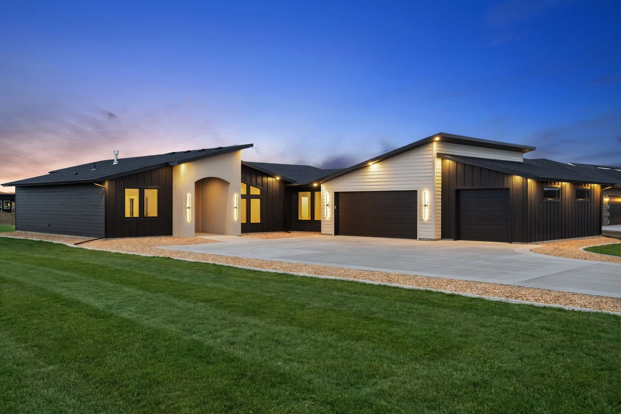 Modern house with black and white exterior, illuminated by outdoor lighting at dusk, with a well-manicured lawn and a concrete driveway.