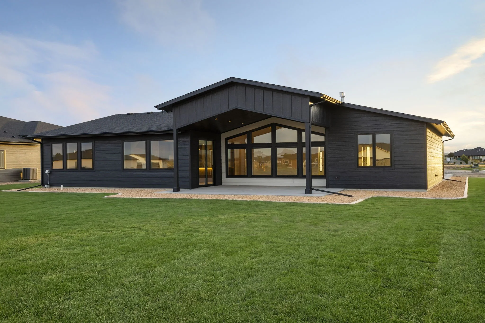 Backyard view of a modern house with black siding, large windows, a lawn, and a gravel pathway, under a clear sky.