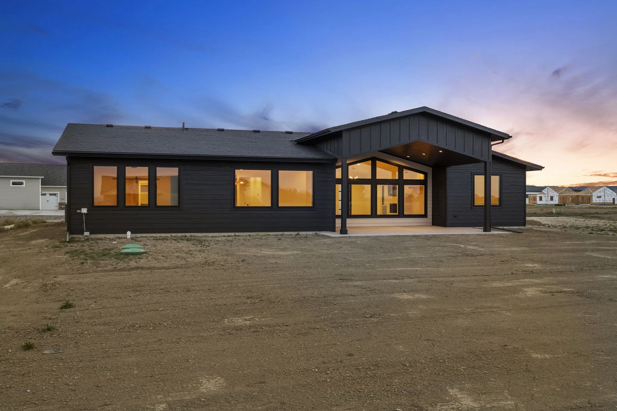 New dark gray house with large windows, illuminated interior, and a covered porch, under a sunset sky with neighboring houses under construction in the background.