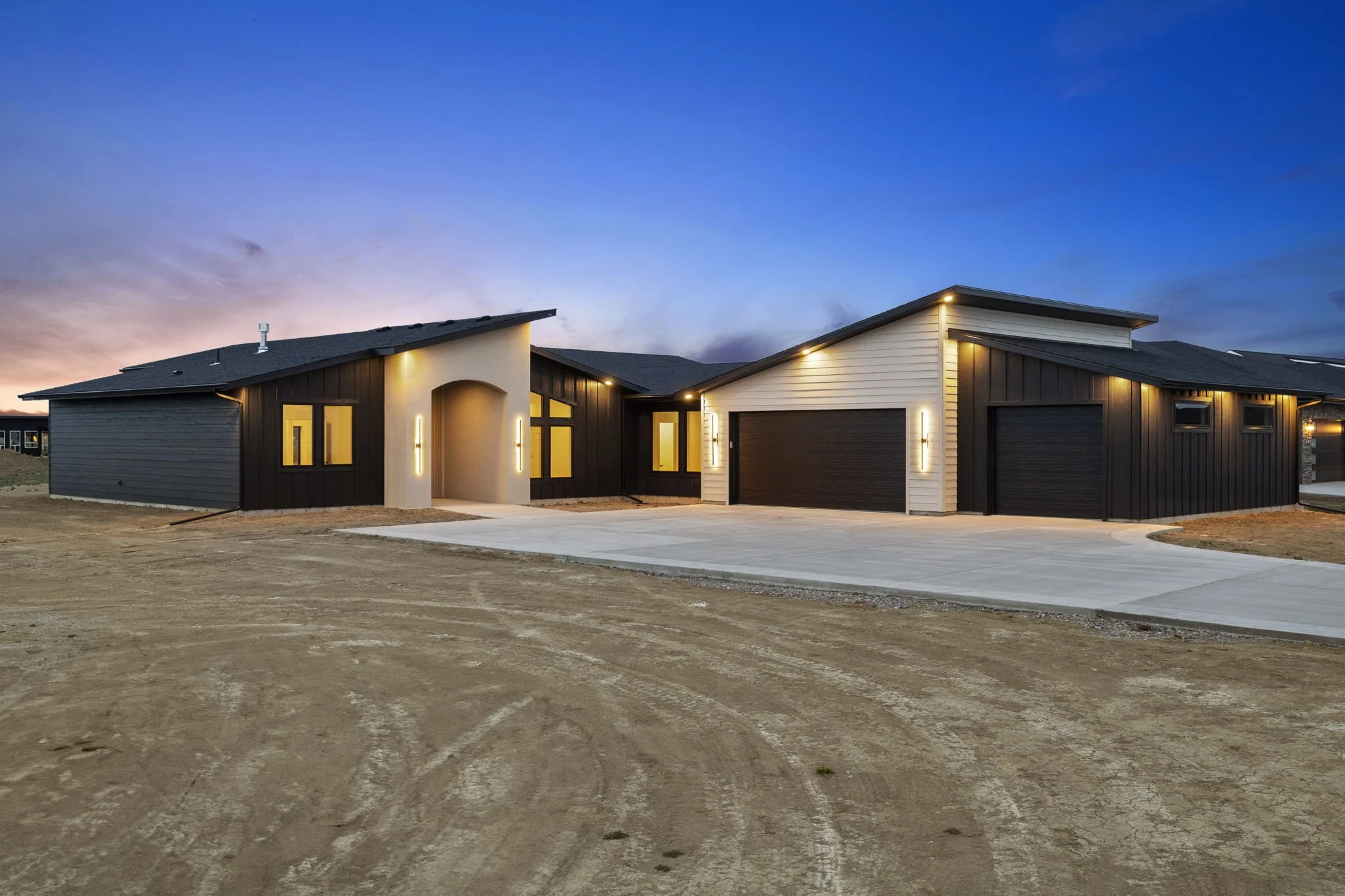 Modern house with black and white exterior, large windows, front driveway, and minimal landscaping, sunset sky in the background.