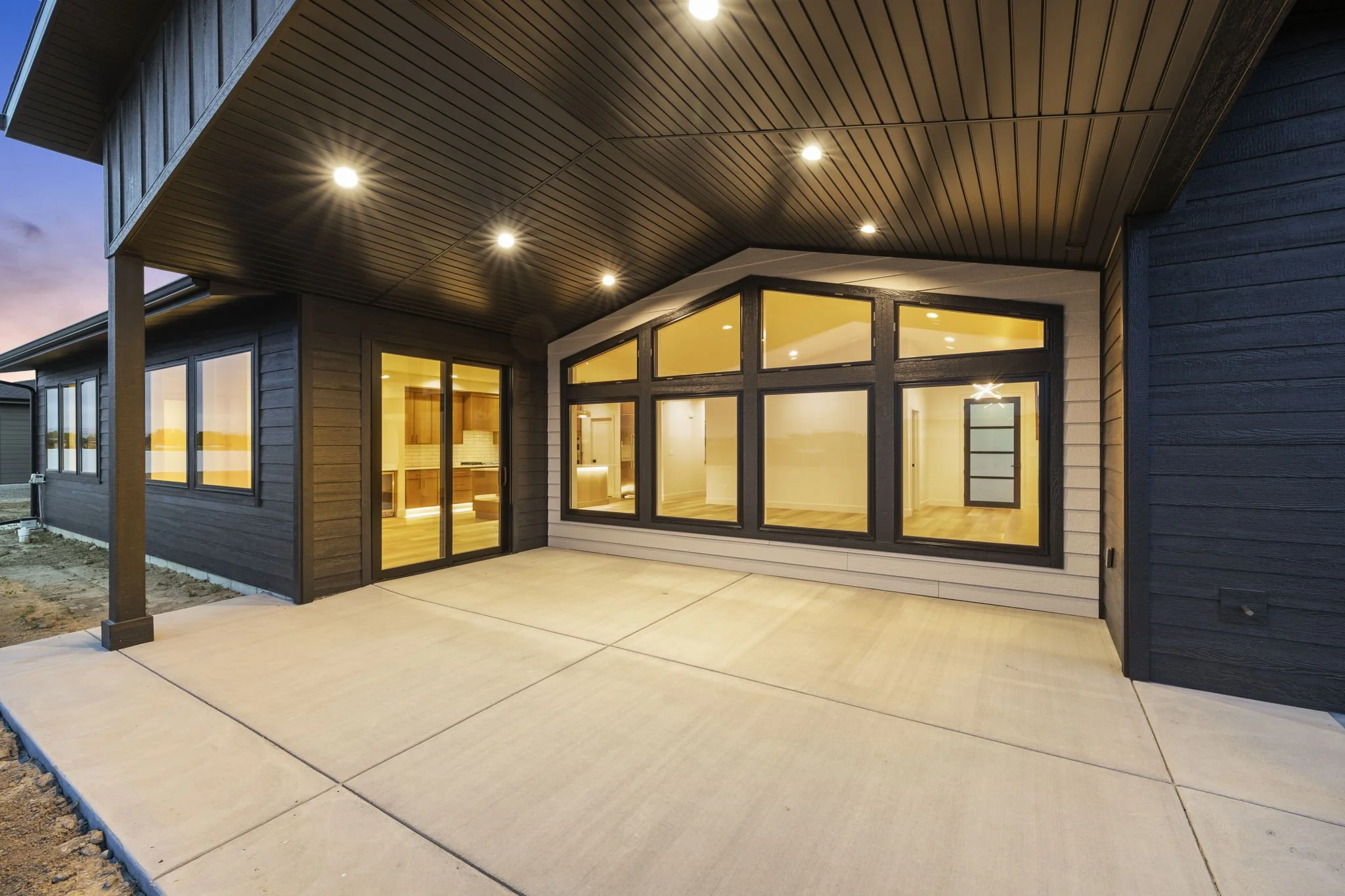 Modern house with a large covered patio, black window frames, and interior lighting visible through glass doors and windows.