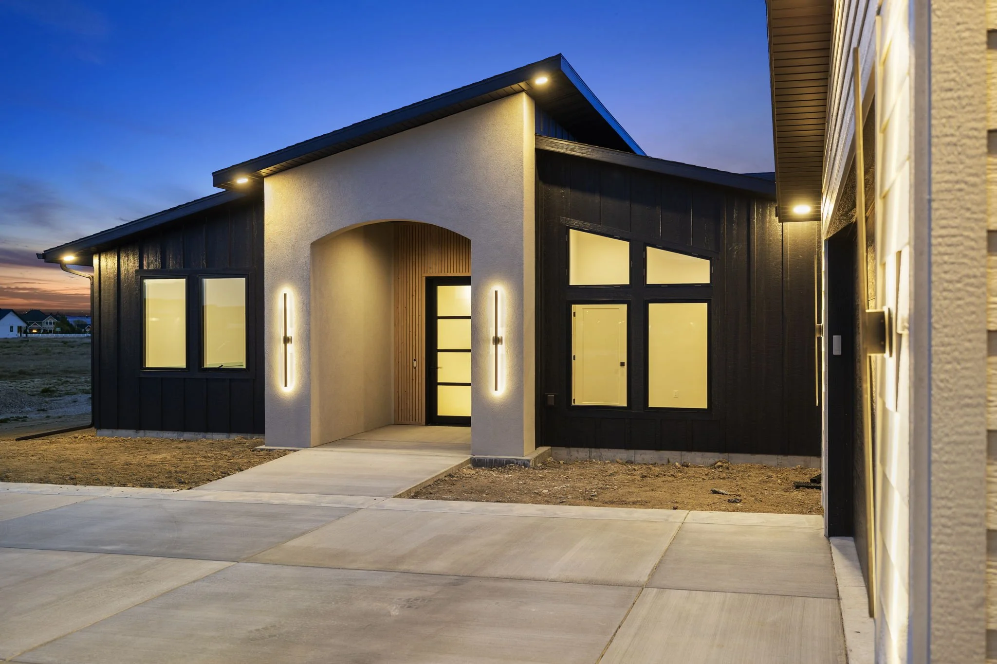 Modern house with black and white exterior, large windows, and warm interior lighting during dusk, with a concrete walkway leading to the front door.