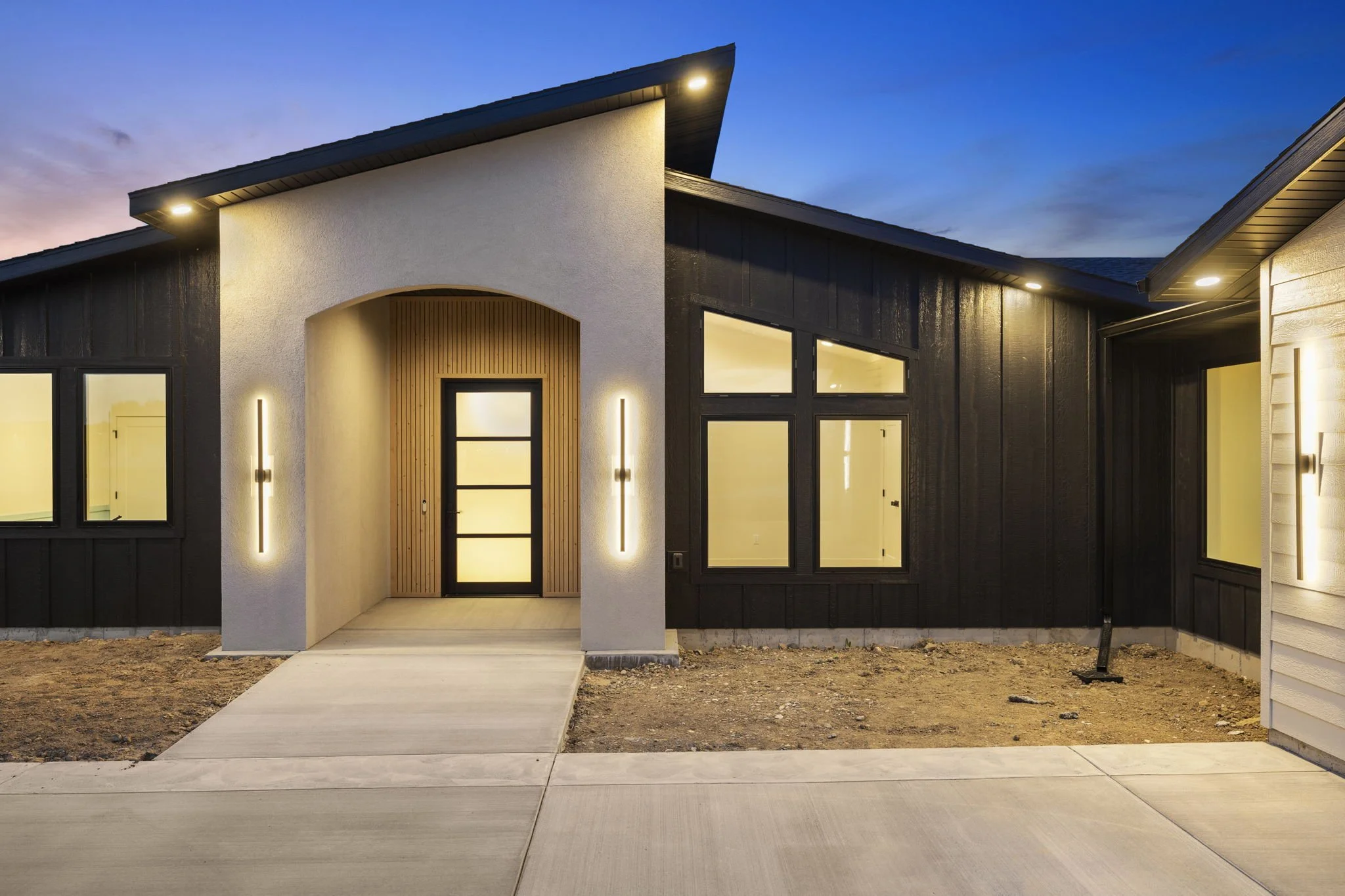 Modern house with black and white exterior, lit front entrance, and large windows at dusk.