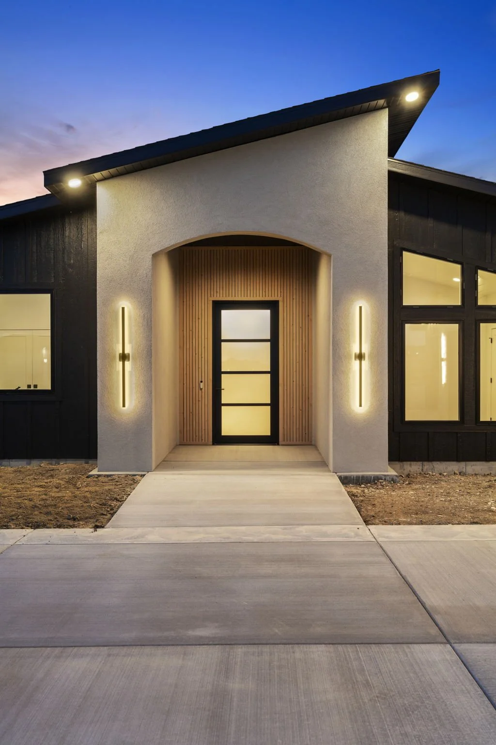 Modern house entrance with a black door, wood paneling, and vertical lights on a white textured wall at dusk.