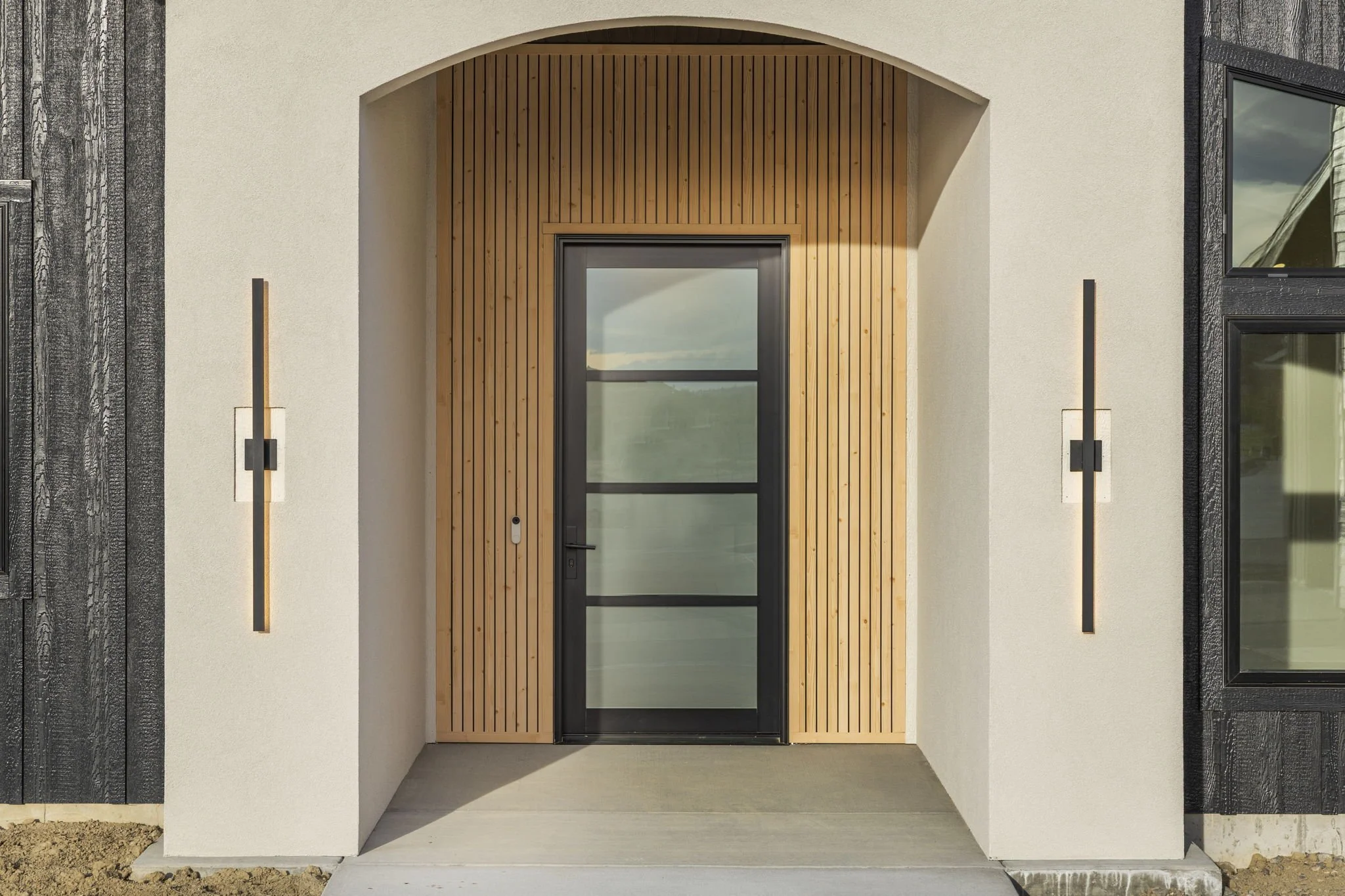 Modern house entrance with a black front door framed by wood paneling and surrounded by white stucco walls, flanked by vertical light fixtures.