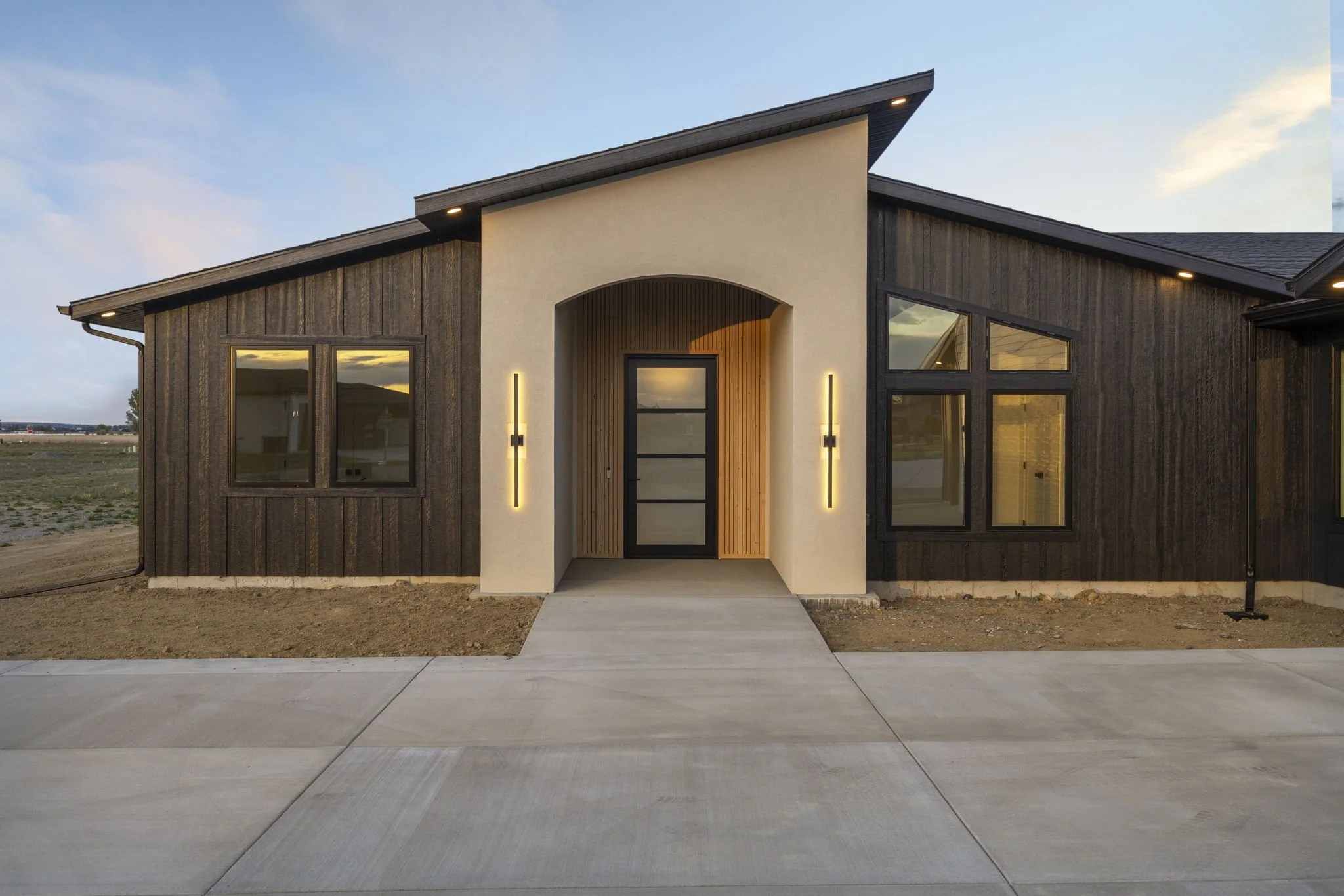 Modern house with a white entrance, black accents, vertical wooden siding, and large windows, under a partly cloudy sky during sunset.