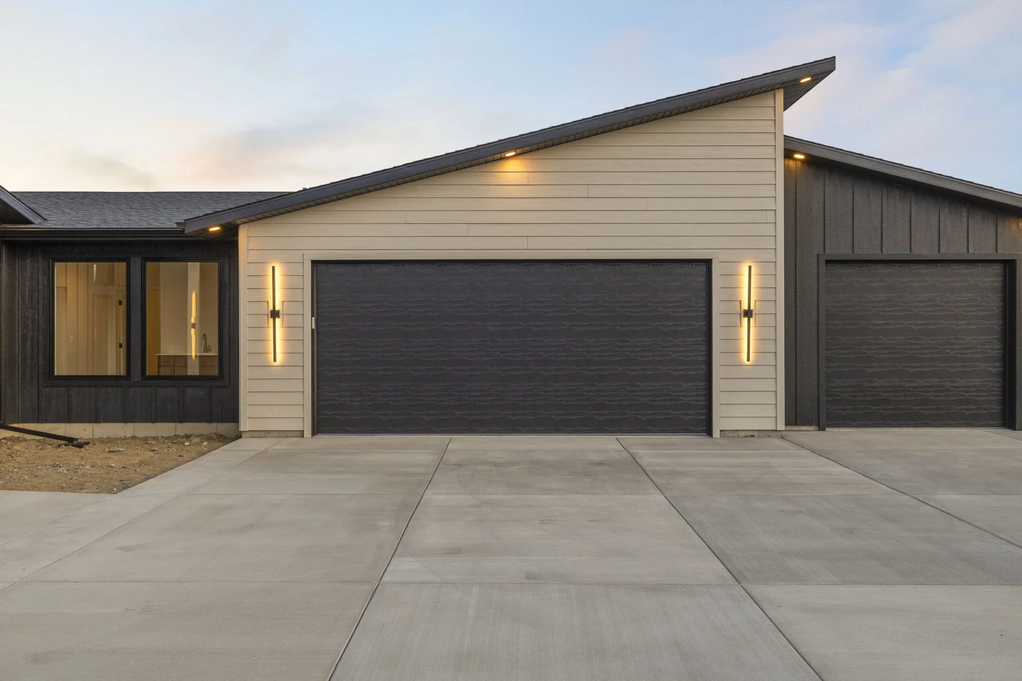 Modern residential house with a two-car garage, light-colored siding, dark garage doors, and minimal outdoor lighting at dusk.