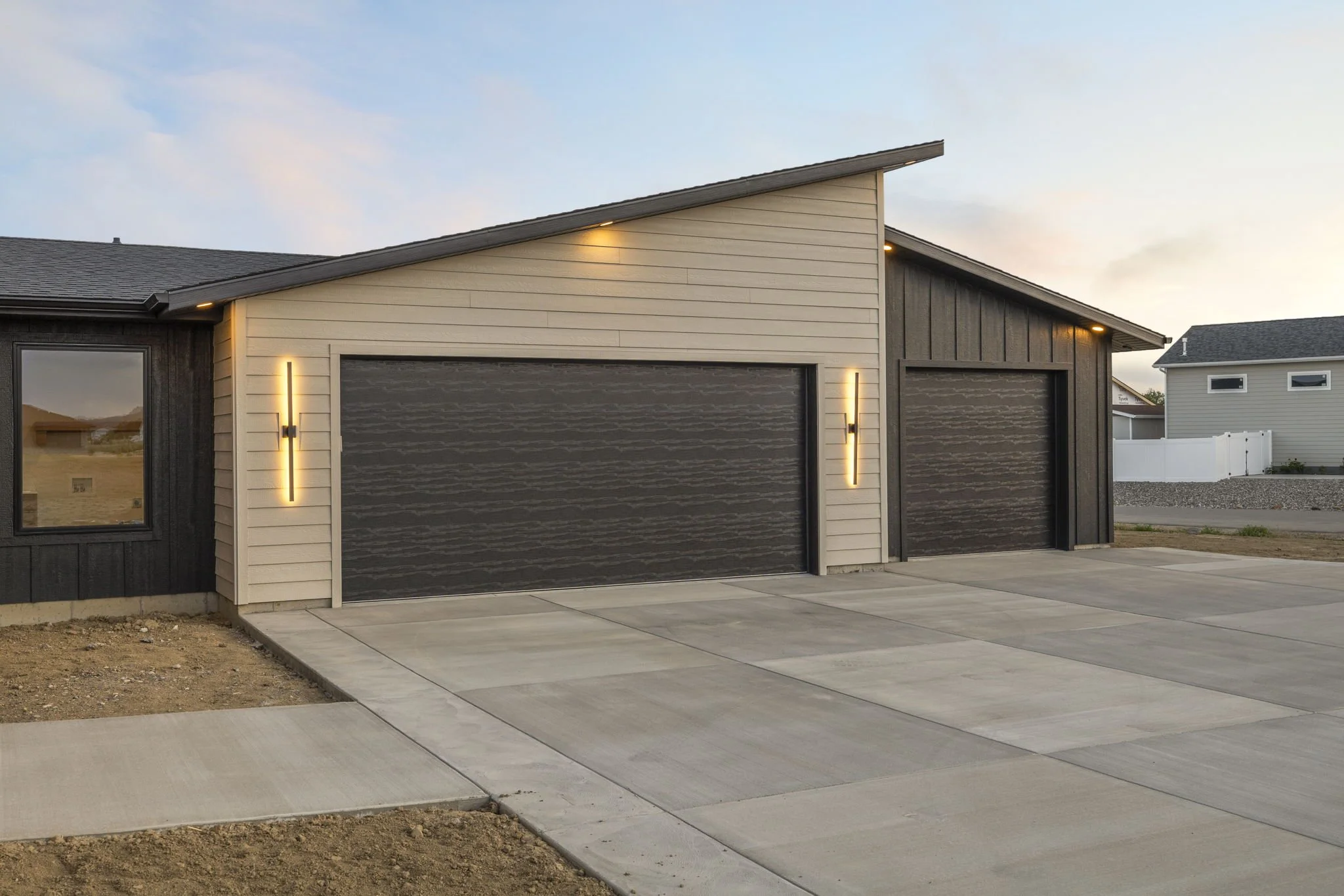 A modern house with two garage doors, exterior lighting, and a concrete driveway and sidewalk in the evening or early morning.