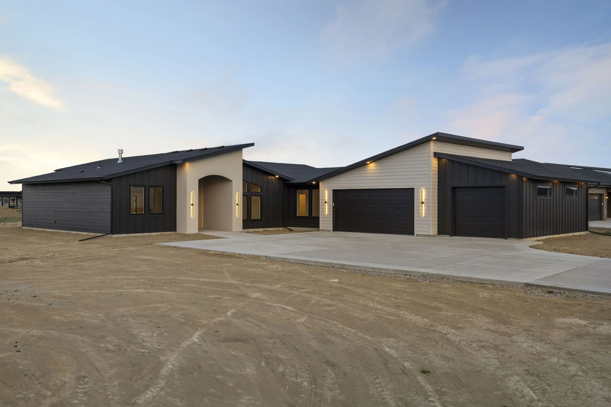 Modern house with black and white exterior, garage door, and outdoor lighting, situated on a dirt lot with a cloudy sky above.