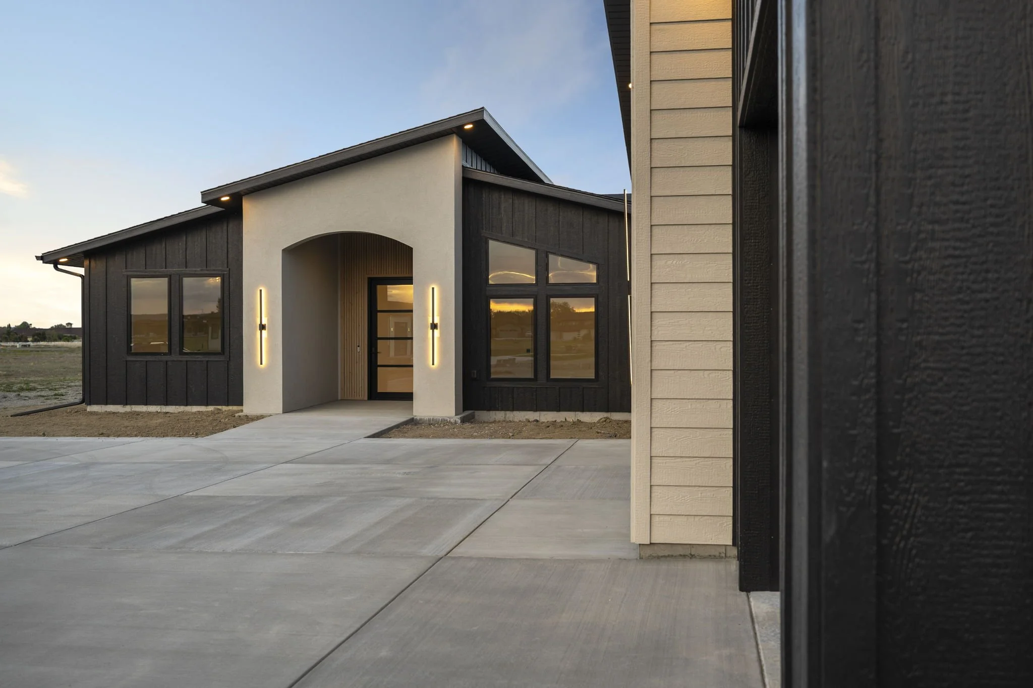 Modern house with a beige and black exterior, front entrance framed by vertical wood paneling, and large windows reflecting a sunset sky, with a concrete driveway in the foreground.