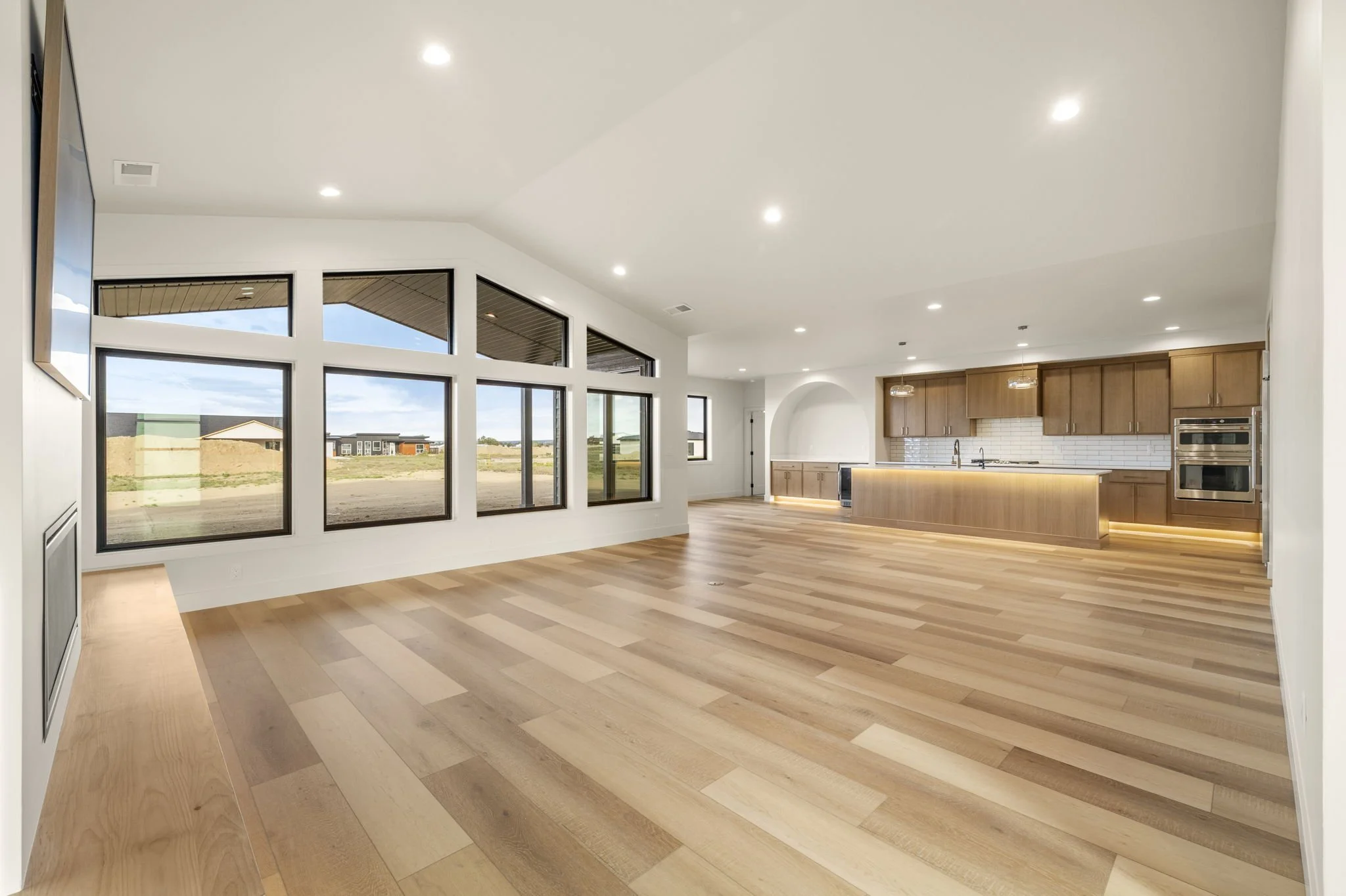 Empty modern living space with large windows, hardwood flooring, and a kitchen area with wooden cabinets and white tile backsplash.
