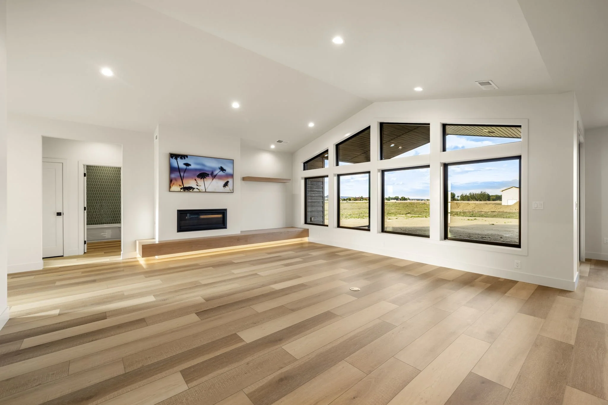 Bright living room with large windows showing an outdoor view, hardwood floors, a wall-mounted flat-screen TV, and a modern fireplace.