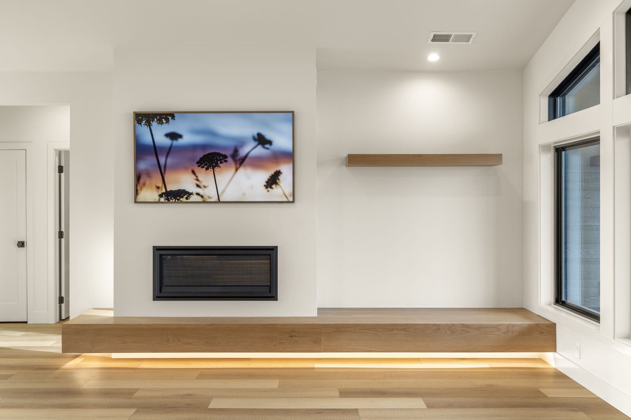 Interior of a modern living room with white walls, wooden flooring, a large flat-screen TV displaying a silhouette of flowers against a sunset sky, and a window on the right side.