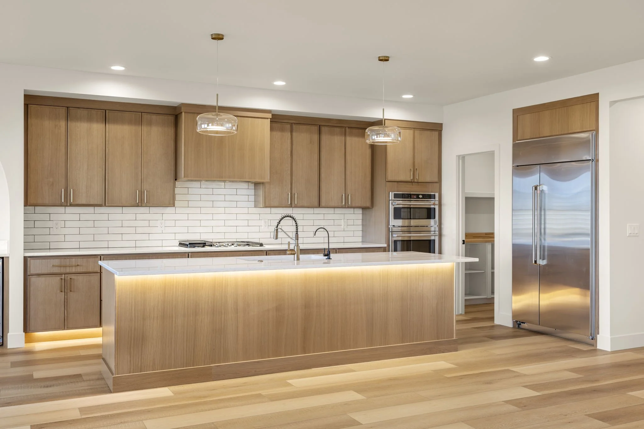Modern kitchen with wood cabinets, a white island with lighting below, a white subway tile backsplash, built-in stainless steel oven, and a stainless steel refrigerator.