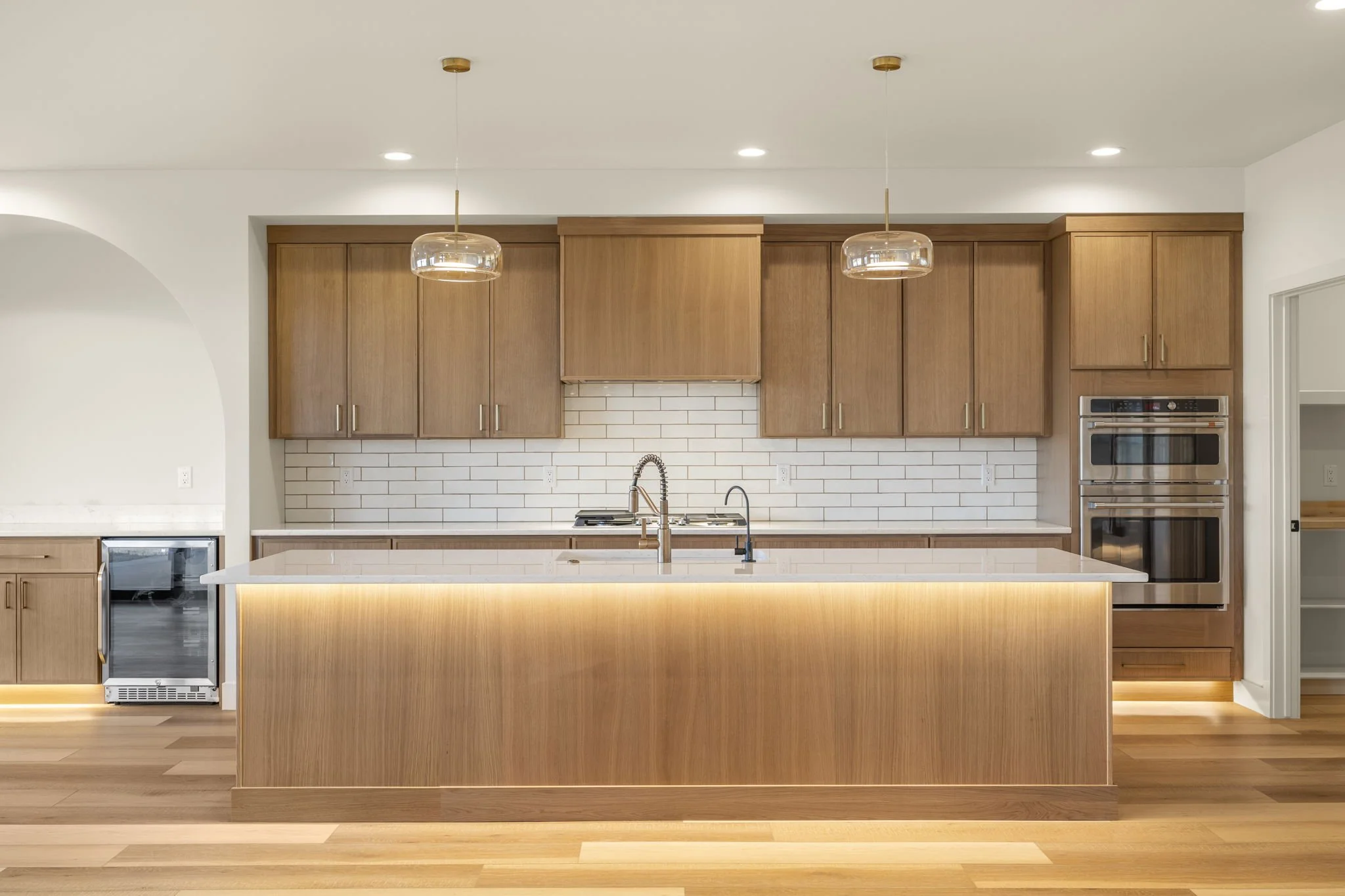 Modern kitchen with wooden cabinets, a white island with a sink and a gold faucet, two pendant lights, white subway tile backsplash, built-in oven and microwave, and hardwood floors.