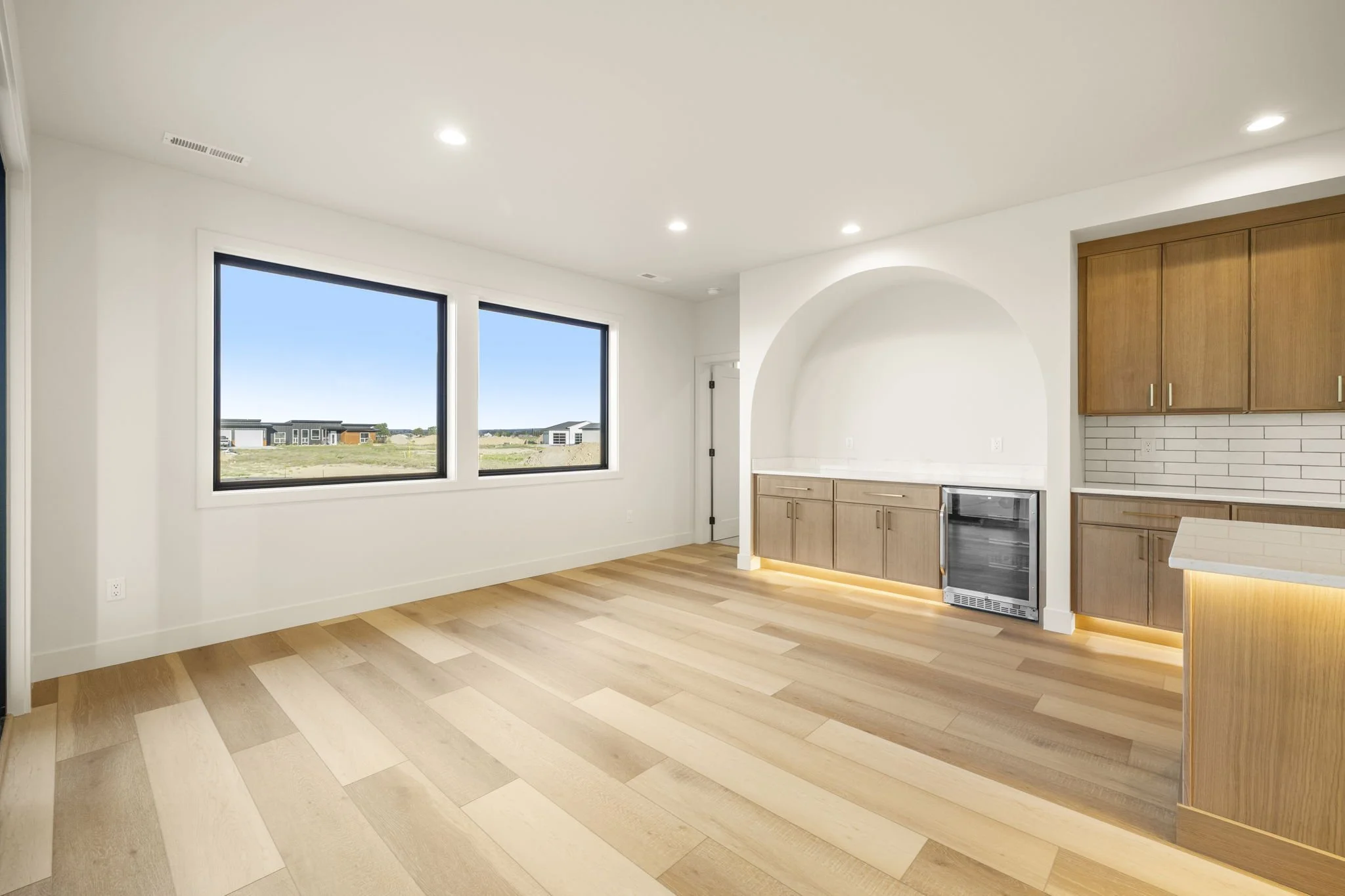 Empty modern kitchen with large windows, wooden cabinets, light hardwood floors, and a view of neighboring houses outside.