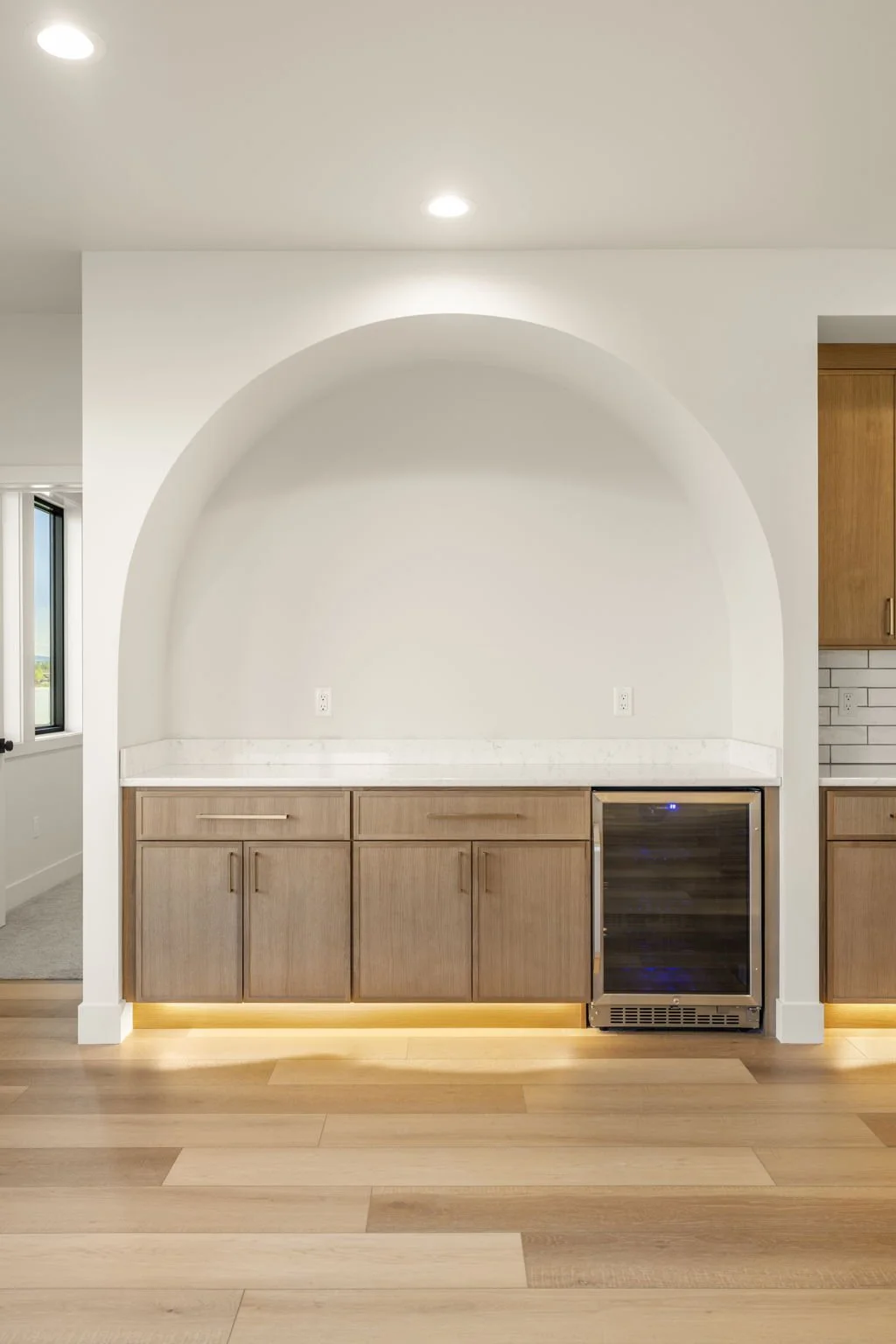Modern kitchen with wooden cabinets, a small wine fridge, and white walls with a decorative arch and under-cabinet lighting.
