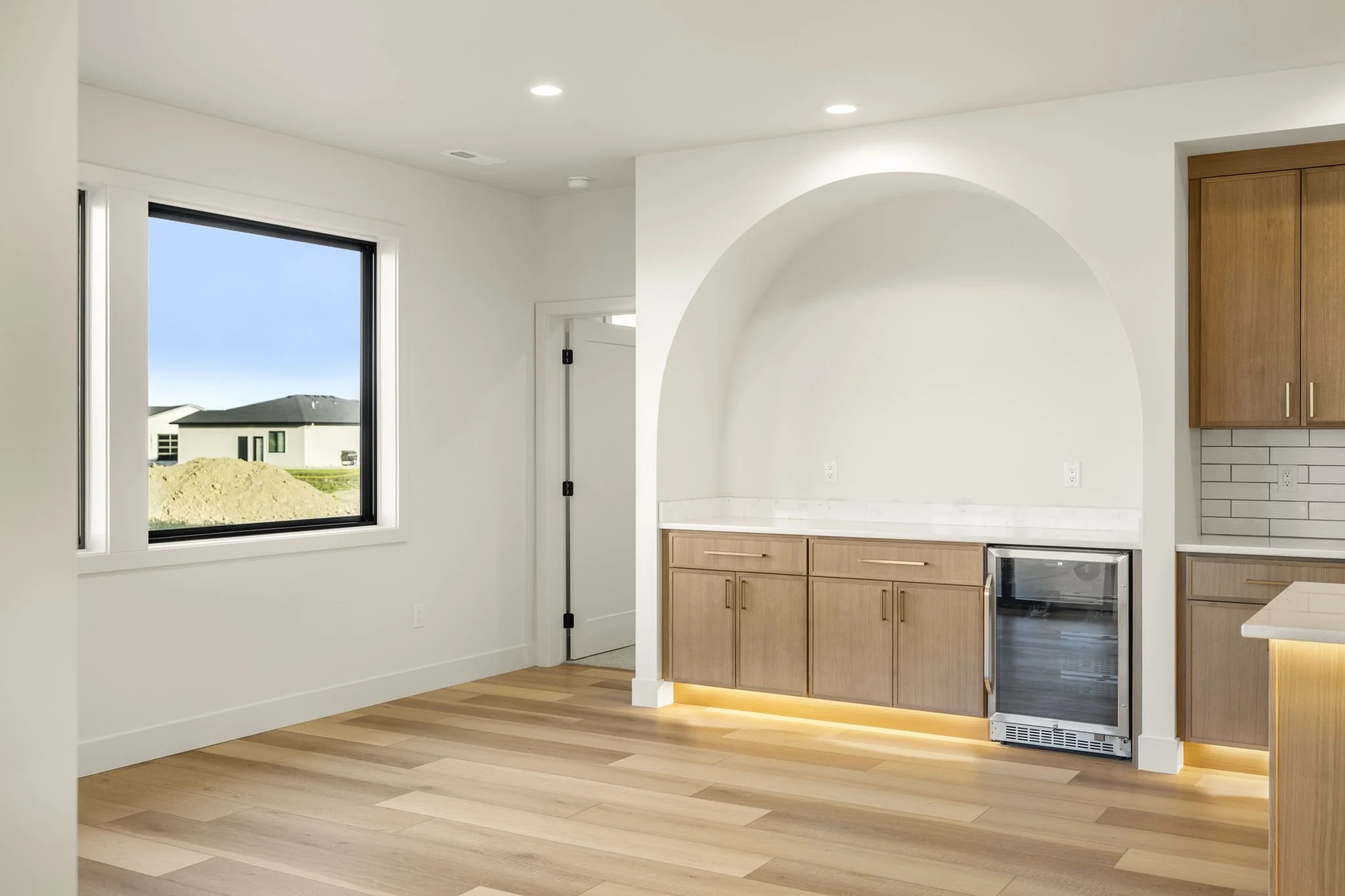 Interior view of a modern kitchen with white walls, wooden cabinets, a small stainless steel refrigerator, a window showing a house and yard outside, and recessed ceiling lights.