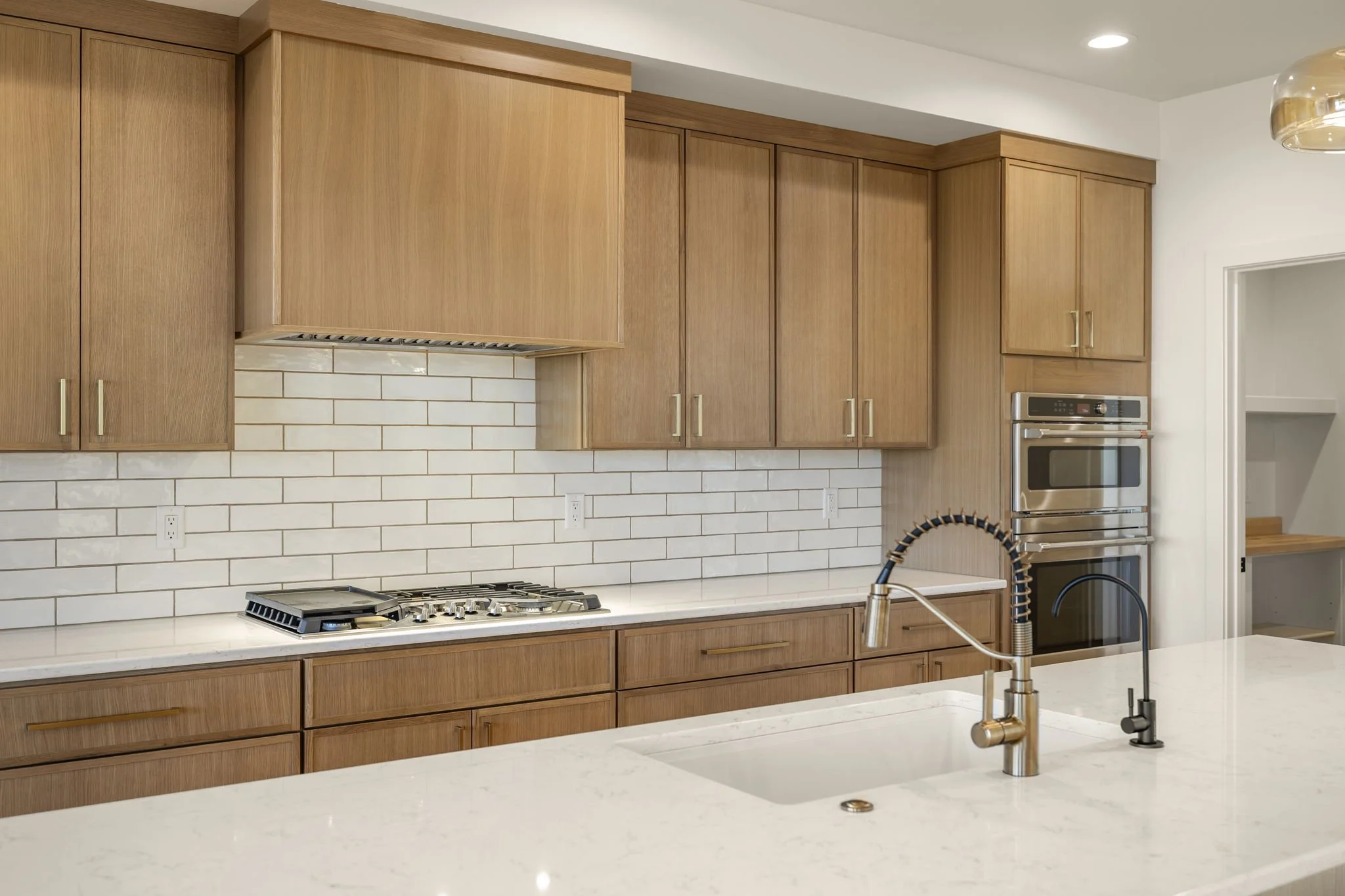 Modern kitchen with wooden cabinets, white subway tile backsplash, stainless steel double oven, gas stove, white marble countertop, and a stainless steel faucet with a black pull-down sprayer.