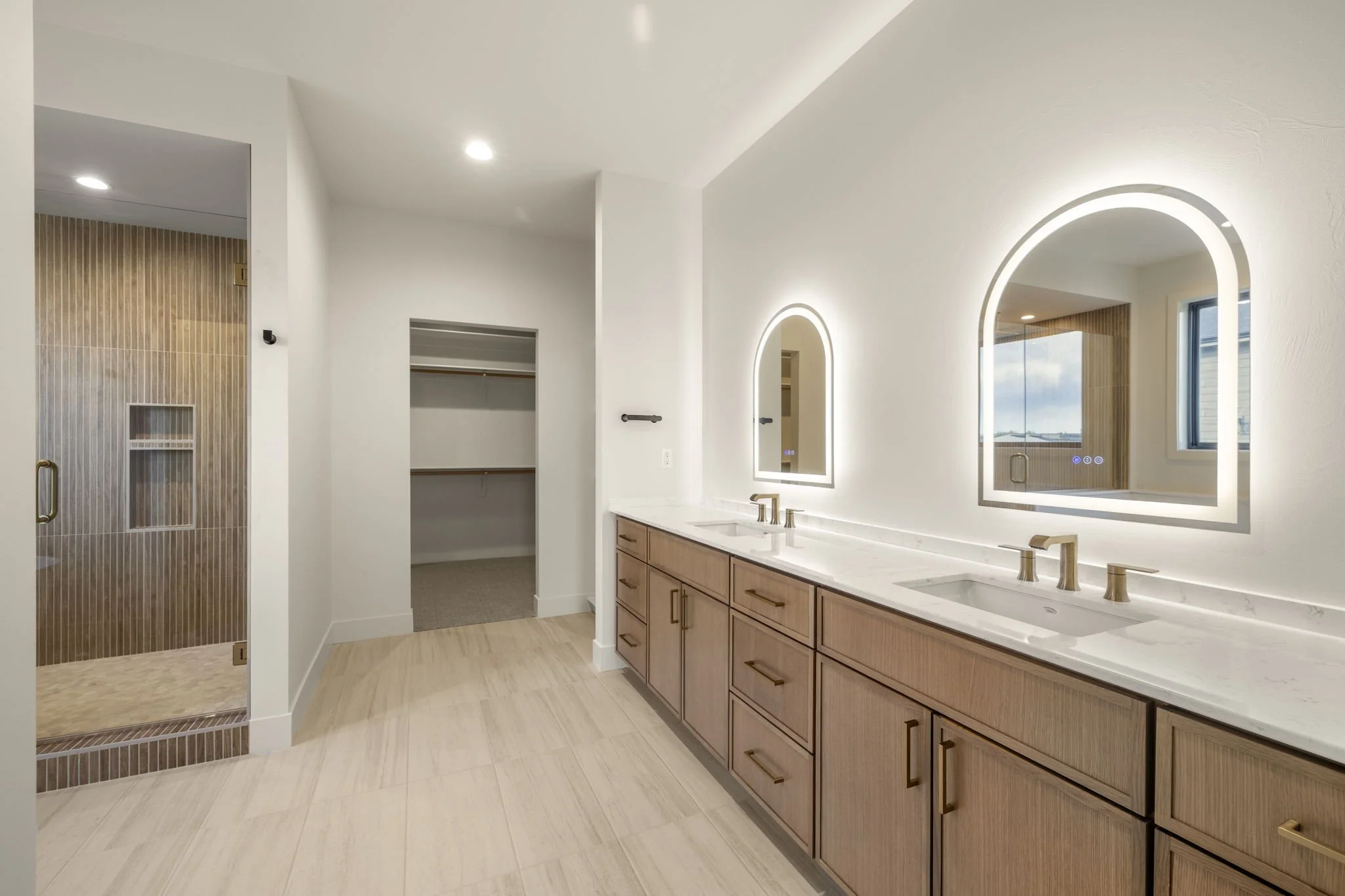 Modern bathroom with double sinks, wooden cabinets, large illuminated mirrors, light-colored flooring, and a walk-in shower with a wooden door and glass panel.