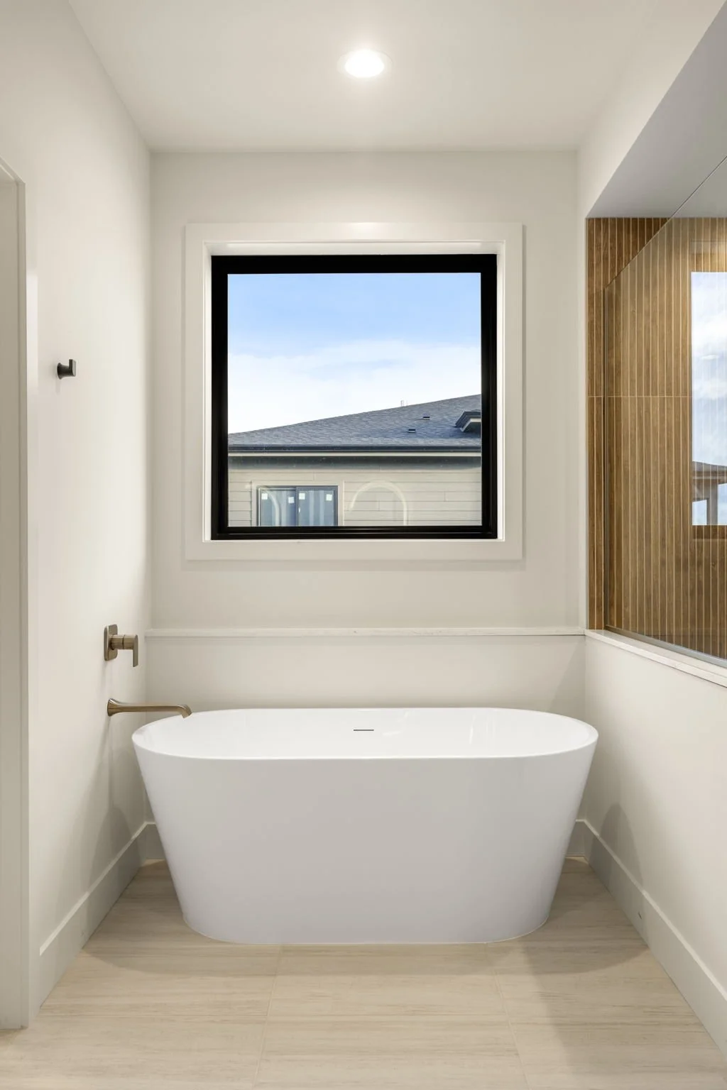 Modern bathroom with a white freestanding bathtub, window with a view of neighboring houses, and a wooden-accented wall.