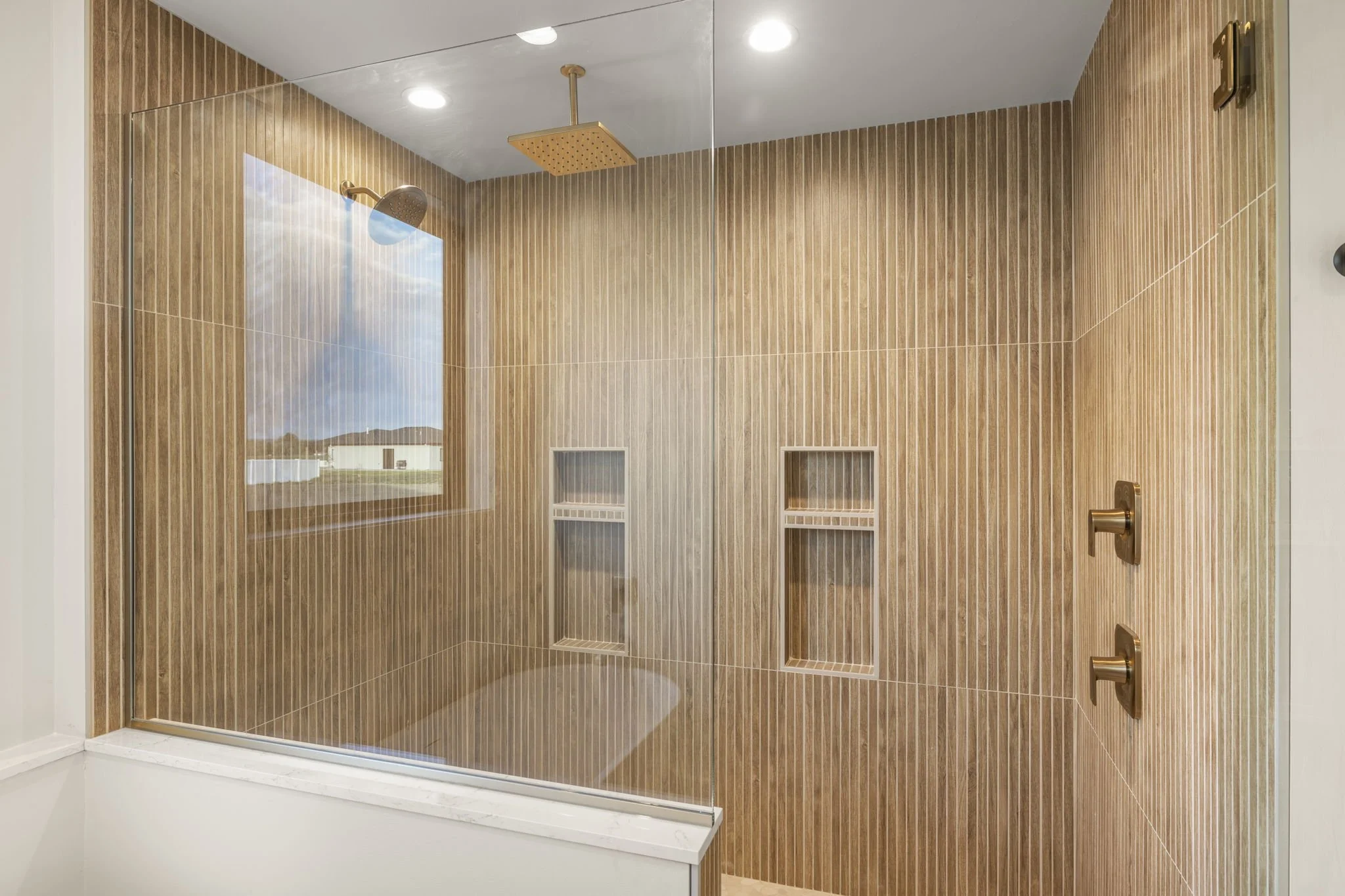 A modern shower with a glass enclosure, wooden vertical tile walls, a square rain shower head, a handheld shower head, and built-in niche shelves, with a window showing a blue sky and hills outside.