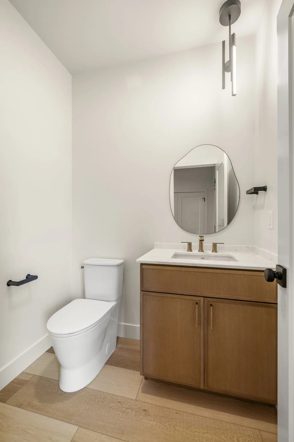A minimalist bathroom with a white toilet, wooden vanity with brass fixtures, a round mirror, a marble countertop, black towel bar, and a modern ceiling light fixture.