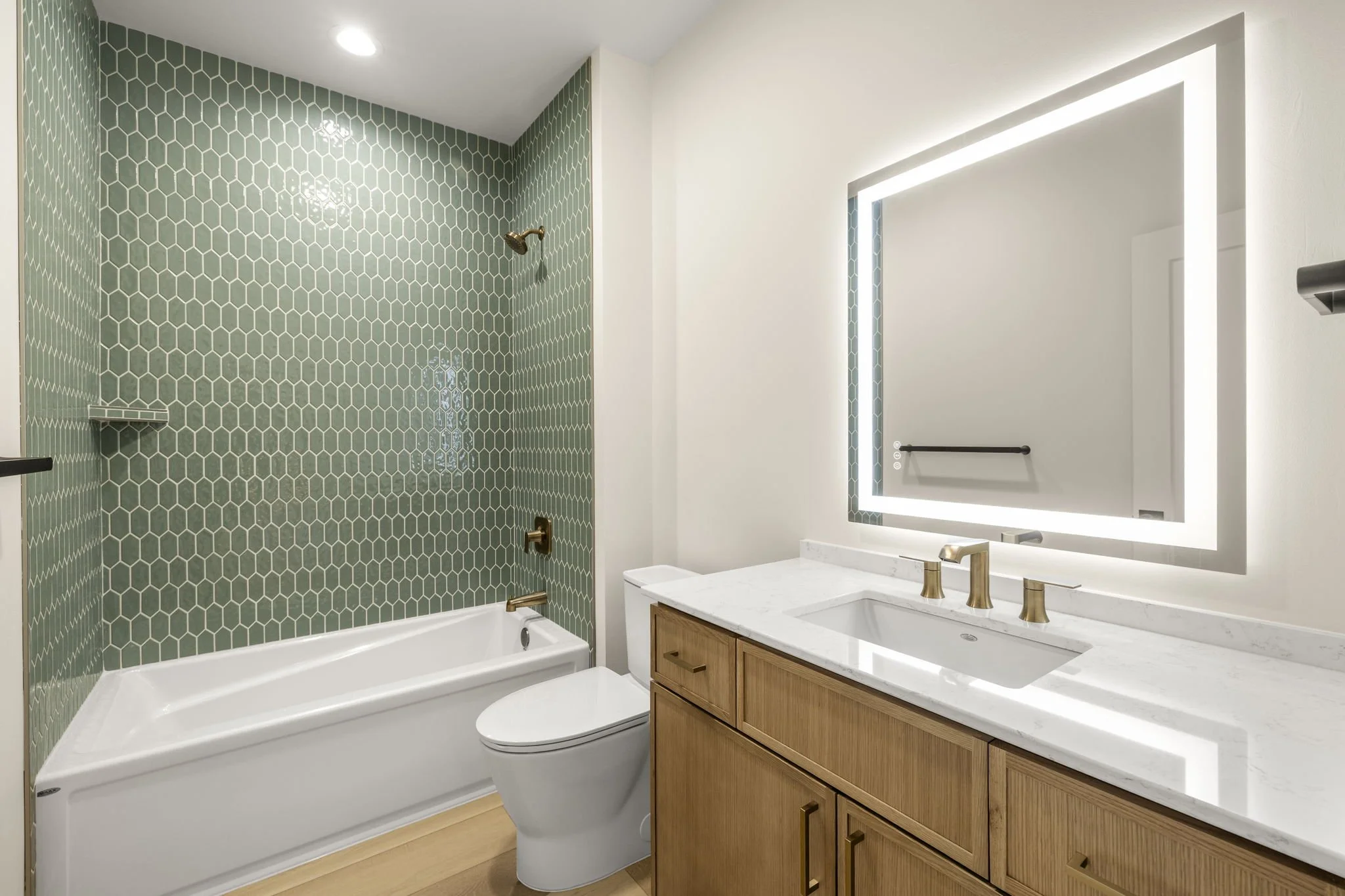 Modern bathroom with a green hexagonal tile shower wall, a white bathtub, a wooden vanity with a white marble countertop, a rectangular mirror with LED lighting, gold fixtures, and light wood flooring.