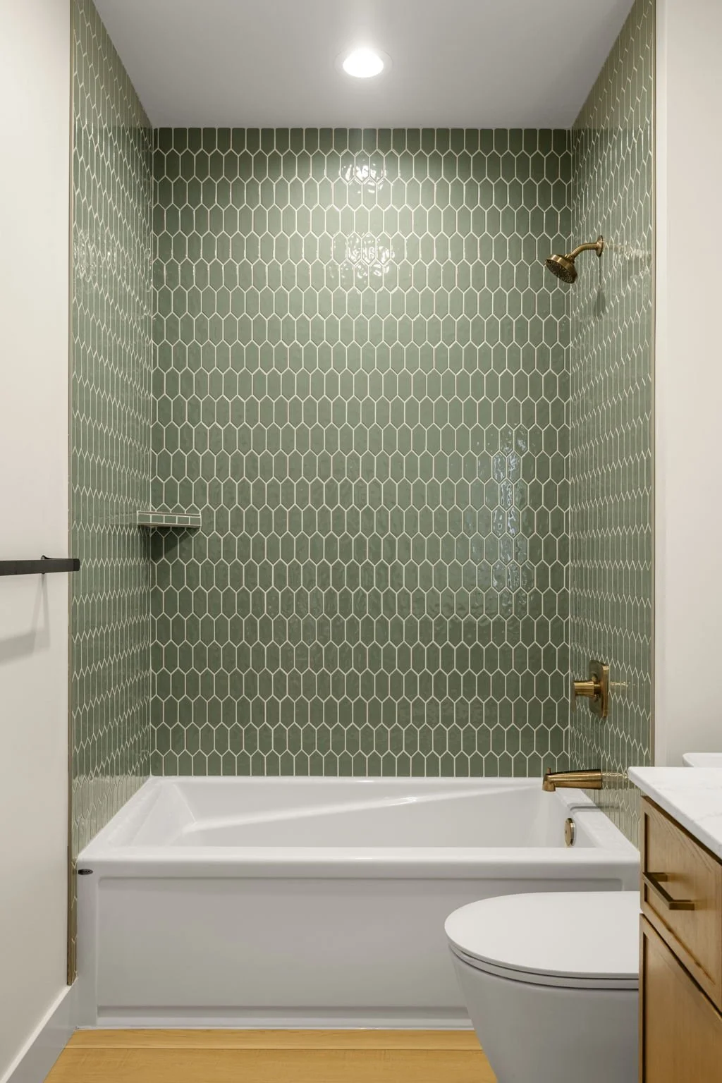 Bathroom with a shower area featuring green hexagonal tiles on the walls, a brass showerhead, a built-in shelf, a white bathtub, a toilet, and a wooden cabinet with a white countertop.