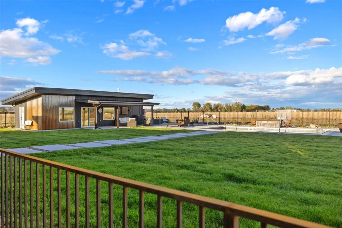 A backyard with a modern house, green lawn, and a swimming pool under a blue sky with clouds.