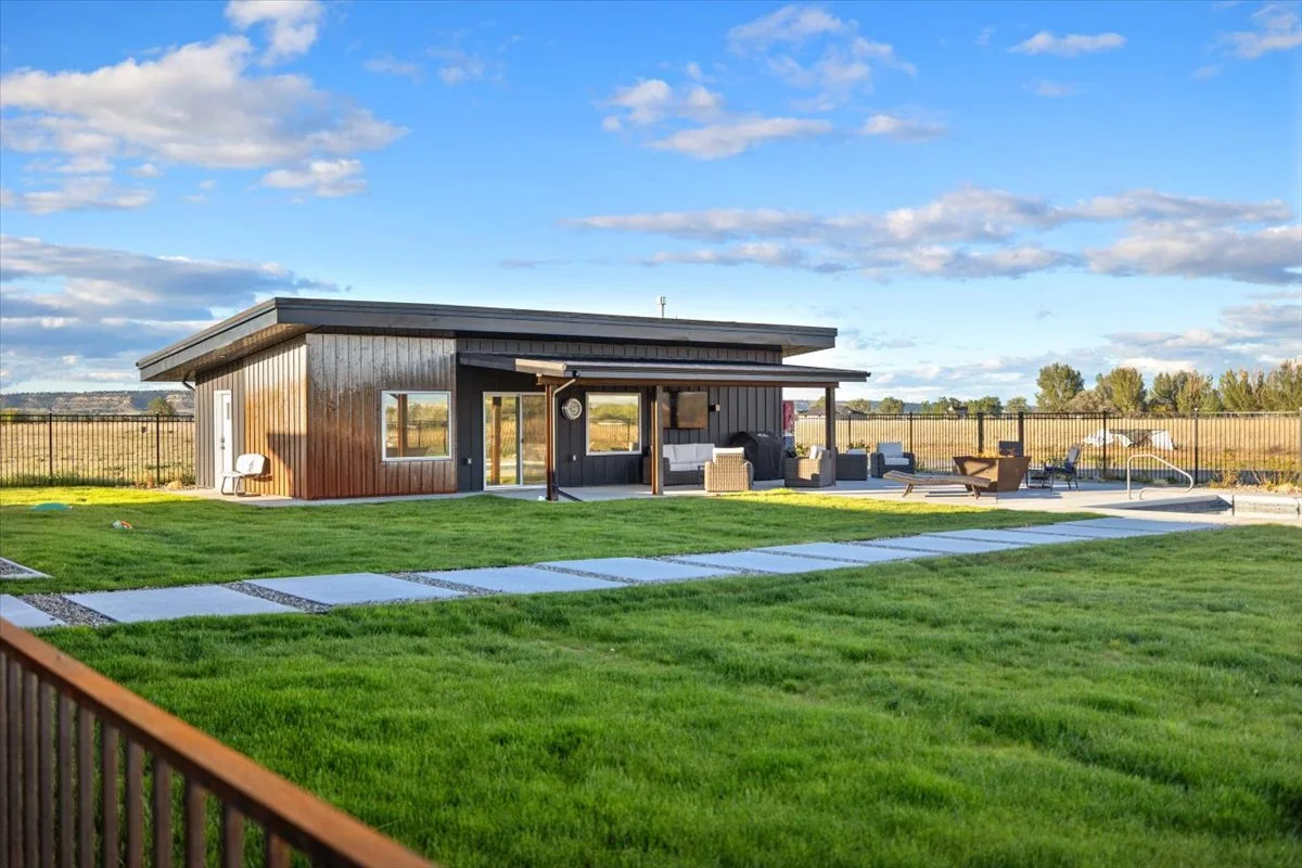A modern backyard with a small wooden house, outdoor seating area, and a grassy lawn under a partly cloudy sky.