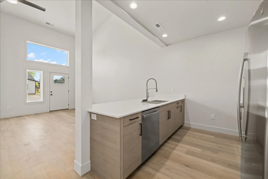 Modern kitchen with light wood cabinets, stainless steel appliances, and white walls, adjacent to a living area with large windows and a front door.