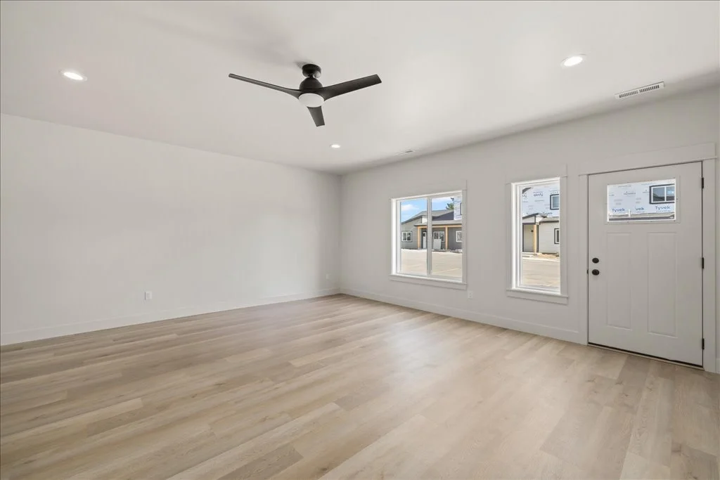 Empty living room with light wood flooring, white walls, two windows, a white front door, and a ceiling fan.