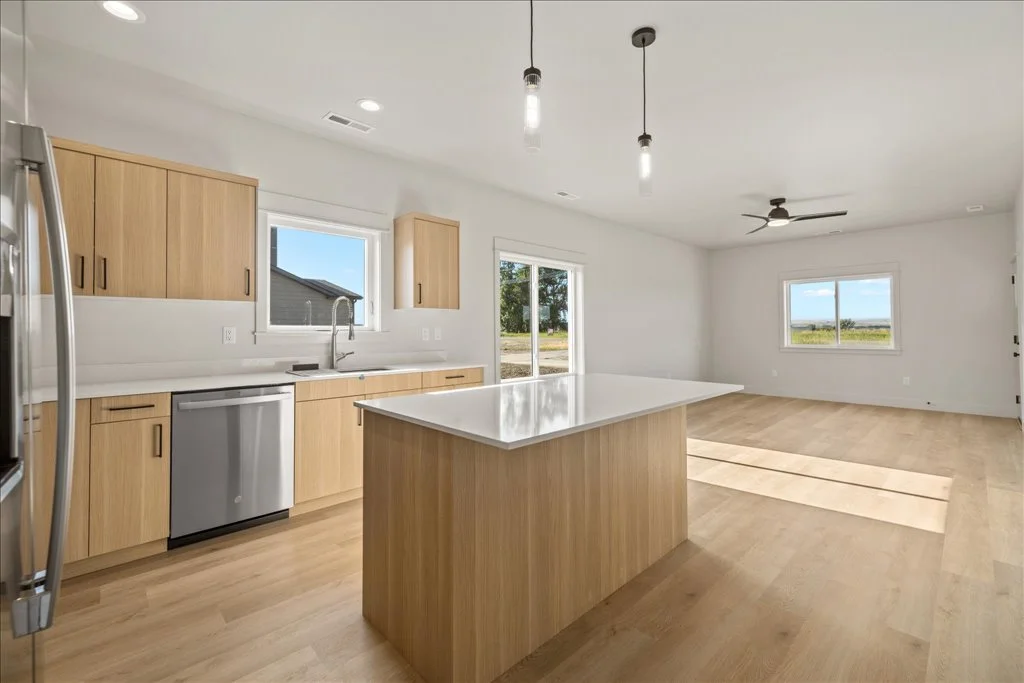 Modern open-concept kitchen and living room with light wooden cabinets, white countertops, stainless steel refrigerator and dishwasher, island, ceiling fans, pendant lighting, and large windows showing a view of the outdoors.