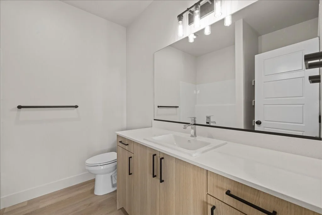 Modern bathroom with a large mirror, white sink, wooden cabinet, and a toilet. Contains black hardware and white walls.