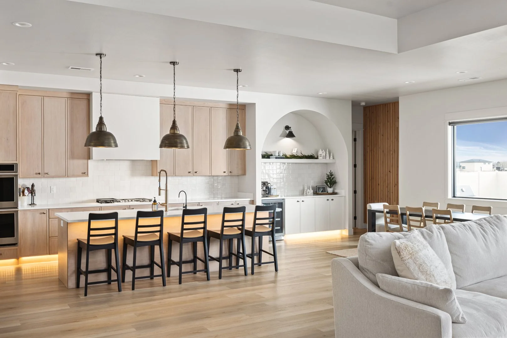 Open kitchen and living area with light wood cabinets, a large island with four chairs, pendant lighting, a sofa, and a dining table near a large window.